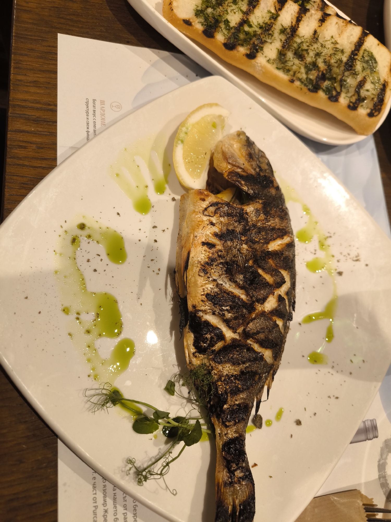 Here is a detailed description of the image:
**Scene:** A top-down view of a restaurant table setting.
**Subjects:** The primary subject is a grilled fish, likely sea bream or similar, garnished with a lemon wedge and small sprigs of green herbs (possibly pea shoots). Next to it is a dish of bread topped with herbs and grill marks. There is a menu card visible.
**Objects:**
* White, square, slightly angled plate holding the fish.
* Grilled fish.
* Lemon wedge.
* Green herbs.
* Oil drizzle on the plate.
* Oblong white dish with bread and herbs.
* Menu card with printed text in a Cyrillic-based language, likely Bulgarian, which translates to "Chardonnay. Rich taste with an elegant structure and fresh finish".
* The table surface appears to be dark wood.
**Actions:** The food is plated and arranged for serving.
**Interactions:** The arrangement indicates that the fish and bread are meant to be eaten together.
**Environment:** The setting suggests a restaurant with a moderately upscale presentation. The lighting is bright, highlighting the food. The table surface appears clean and well-maintained.
**Location:** No specific location in the city of Unknown, Unknown can be identified from the image. The image does not provide any specific information that can point out the location.