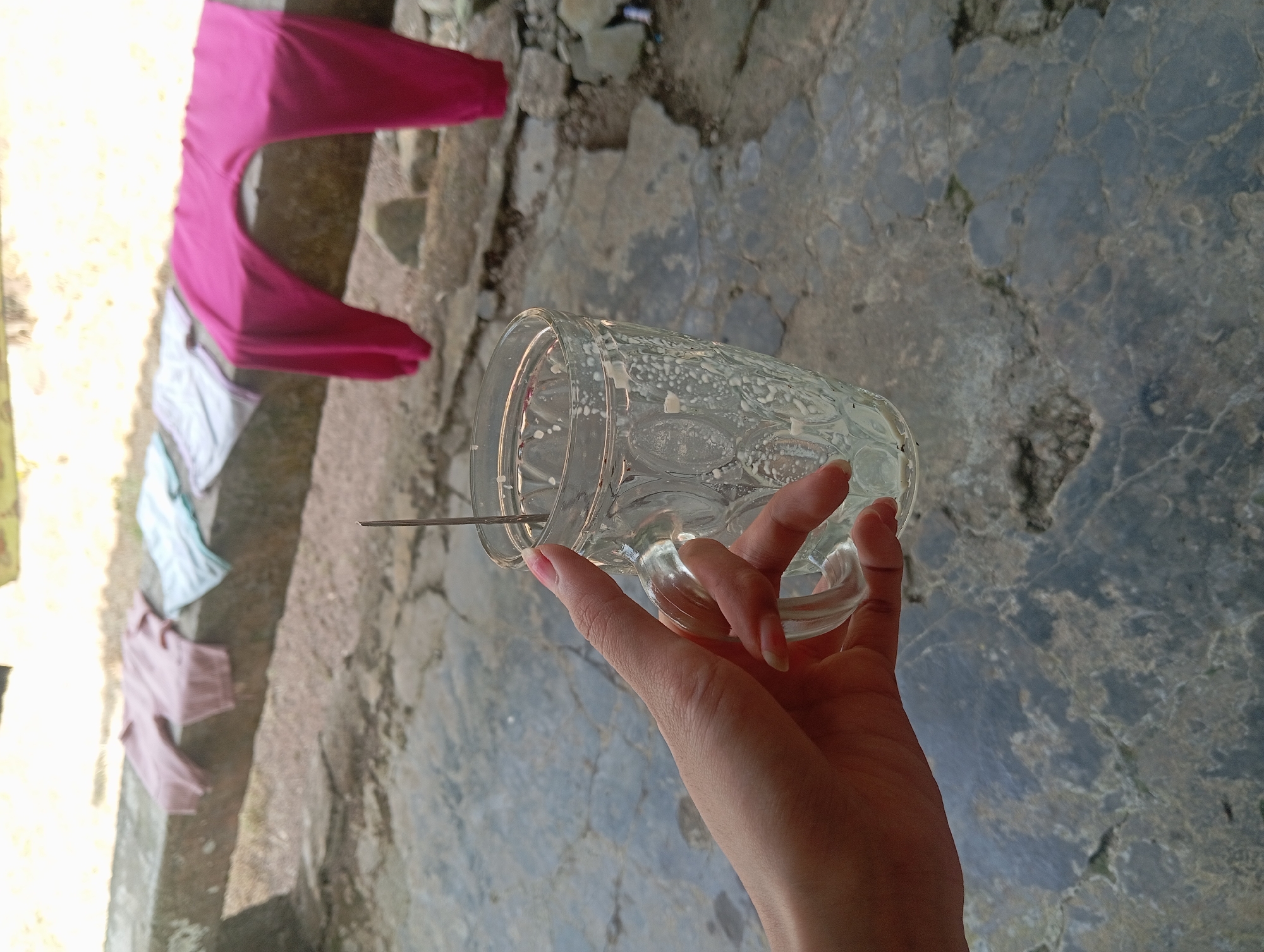 In Rejosari, Indonesia, a person's hand, with fingernails painted light pink, grips a clear glass mug. The mug has a textured, patterned surface and contains a light-colored liquid with visible residue. A thin, metallic object, possibly a stirrer or straw, is partially submerged in the liquid. The background shows clothing laid out on a concrete ledge; specifically, two pairs of pink trousers and two pieces of lighter colored fabric, possibly shirts or shorts. The ground is paved with irregular, grey stones.
