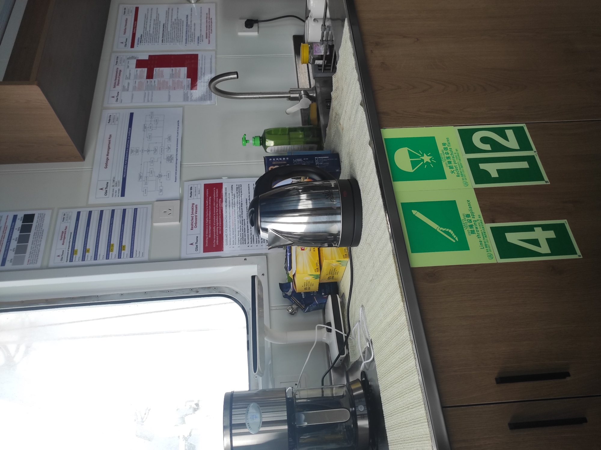 The image shows a galley or pantry area on a ship, likely in the port of Unknown, Unknown. A stainless steel coffee maker and a stainless steel kettle are on the counter, along with boxes of tea. To the right, a stainless steel faucet is mounted over a sink. Various safety posters are visible on the wall, including one about garbage management and another about restricted smoking. Below the counter, there are two cabinets with dark handles. Two green safety signs with white symbols are attached to the cabinets: one depicts a parachute and the other shows a line-throwing appliance. Large white numbers "4" and "12" are displayed within green squares on the cabinets. The scene appears to be clean and well-maintained. There are no people visible in the image.