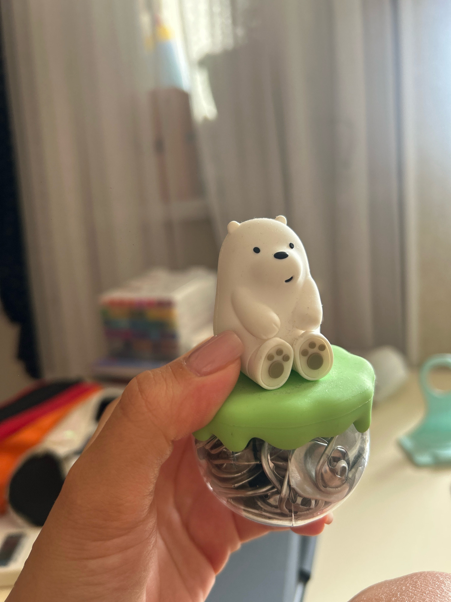 A person's hand, with painted nails, holds a clear plastic container filled with metal thumbtacks, topped with a lime-green silicone lid. A small, white polar bear figurine, seated, rests on the lid. The background is softly blurred, showing a window with curtains, some stacked boxes with colorful edges, and a light-colored surface. The scene is indoors. There is no information available about the location in the city of Khyrdalan, Azerbaijan.
