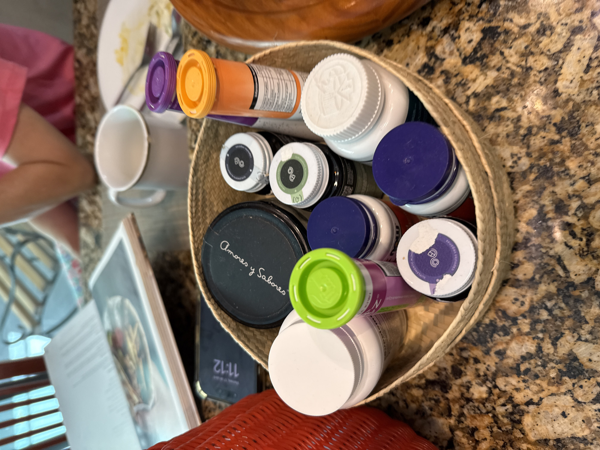 A high-angle indoor shot presents a collection of items in a woven basket on a speckled brown countertop. The basket contains various containers, including pill bottles with purple, orange, green, and white lids. One black lid reads "Amores y Sabores" in white script. A partially visible smartphone screen displays "11:12." Adjacent to the basket, a cookbook rests on a red wicker surface, with a white mug and a plate with food remnants visible in the background. The setting is in San Bernardino Tlaxcalancingo, Mexico. A person wearing a pink shirt is visible on the left side.