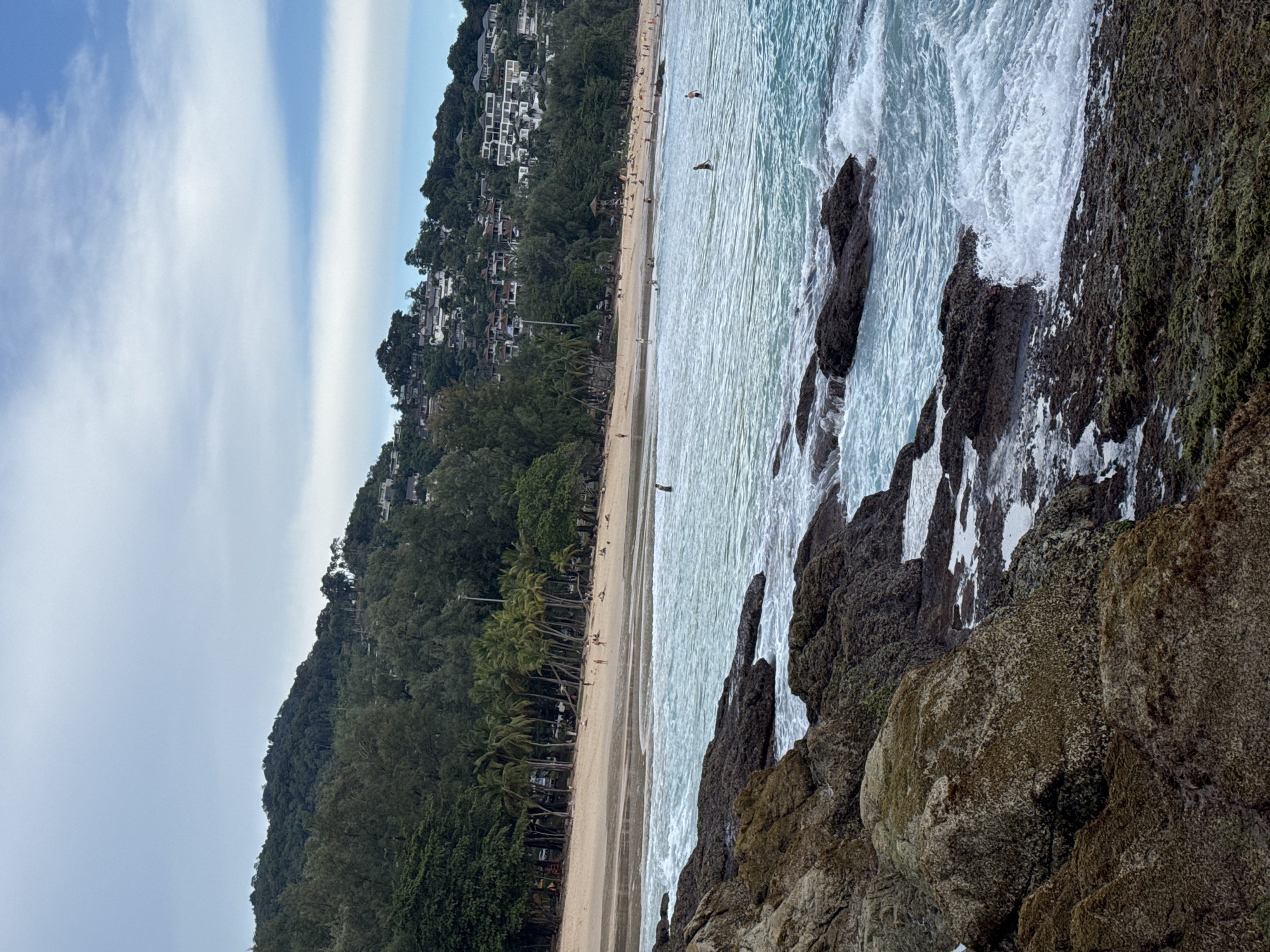 The image depicts a scenic view of a beach in Choeng Thale, Thailand. In the foreground, dark, wet rocks are visible, with waves crashing against them. The ocean water is a clear turquoise blue, with white foam from the waves. A sandy beach stretches across the middle ground, with several palm trees dotting the landscape. In the background, a lush green hillside is covered in dense trees, with houses and buildings nestled among them, suggesting a populated area. The sky is partly cloudy with a mix of blue and white. Several people are visible on the beach and in the water, enjoying leisure activities. The overall impression is a tropical coastal setting, likely a popular tourist destination.