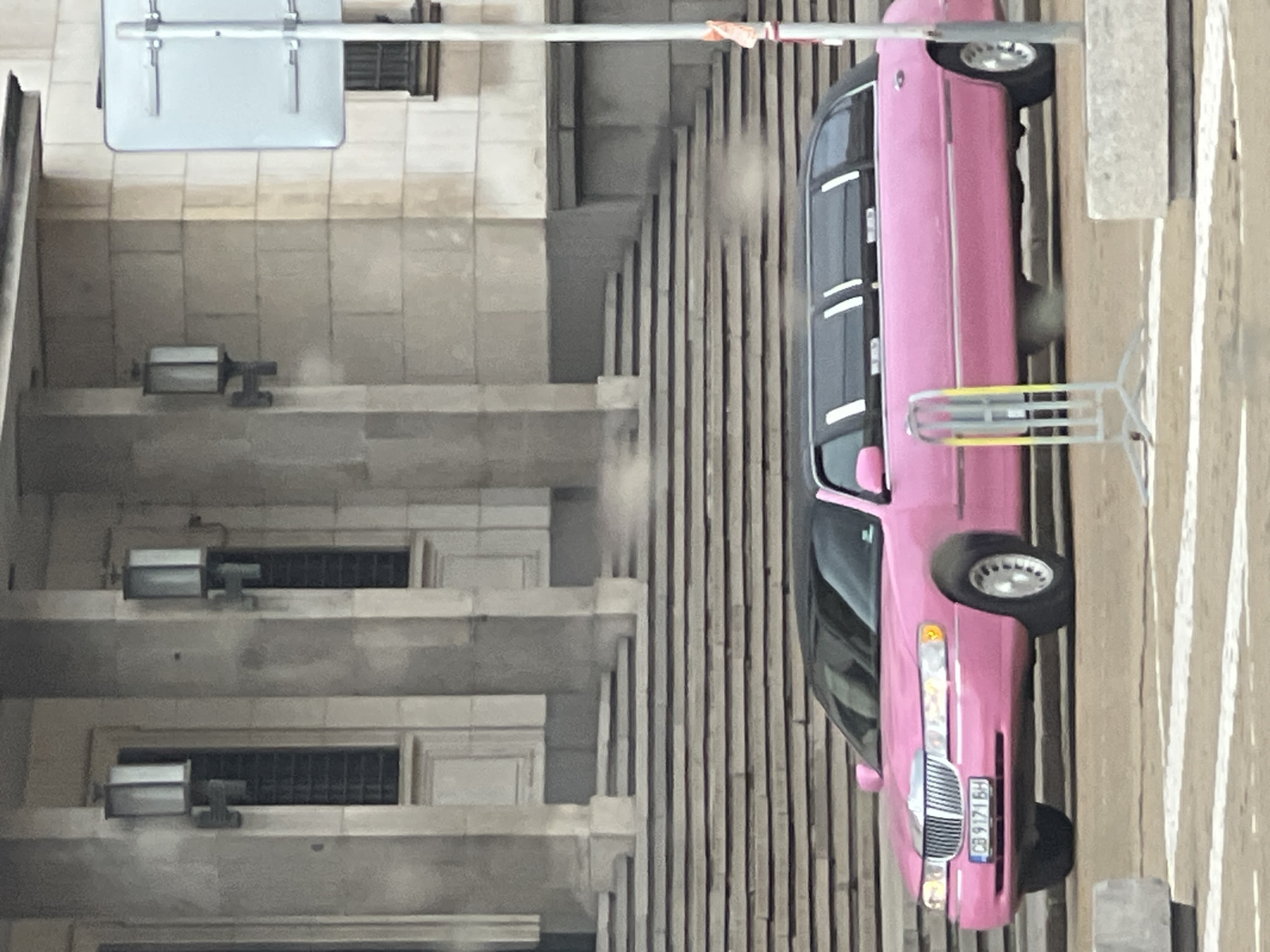 The image shows a pink limousine parked on a street in Sofia, Bulgaria. The license plate is visible and reads "CB 9171 BH". The limousine is next to a building with ornate stone detailing, including windows with metal grates and outdoor lighting fixtures. A pedestrian barrier is next to the limousine on the sidewalk.