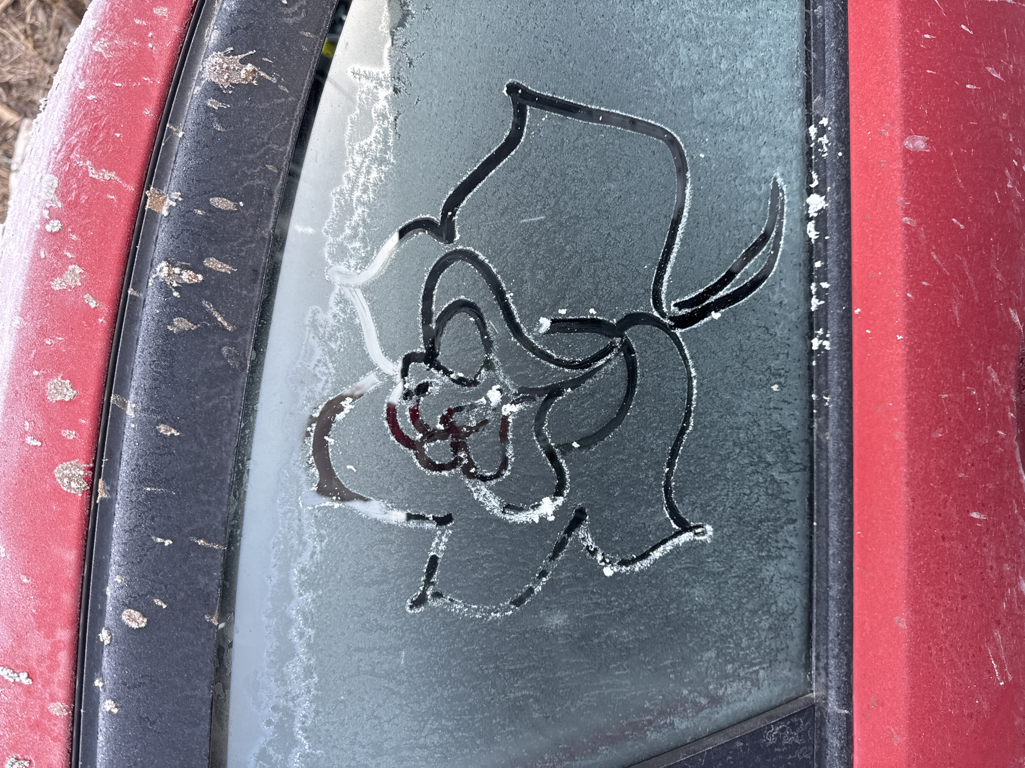 The image shows a red vehicle, likely a car, with a frosted or icy windshield. A design resembling a rose has been traced or etched into the frost on the windshield. The vehicle is dirty with debris, indicating exposure to the elements. The location is not determinable from the image, and there is no information provided to verify the location as Chaikovsky, Russia.