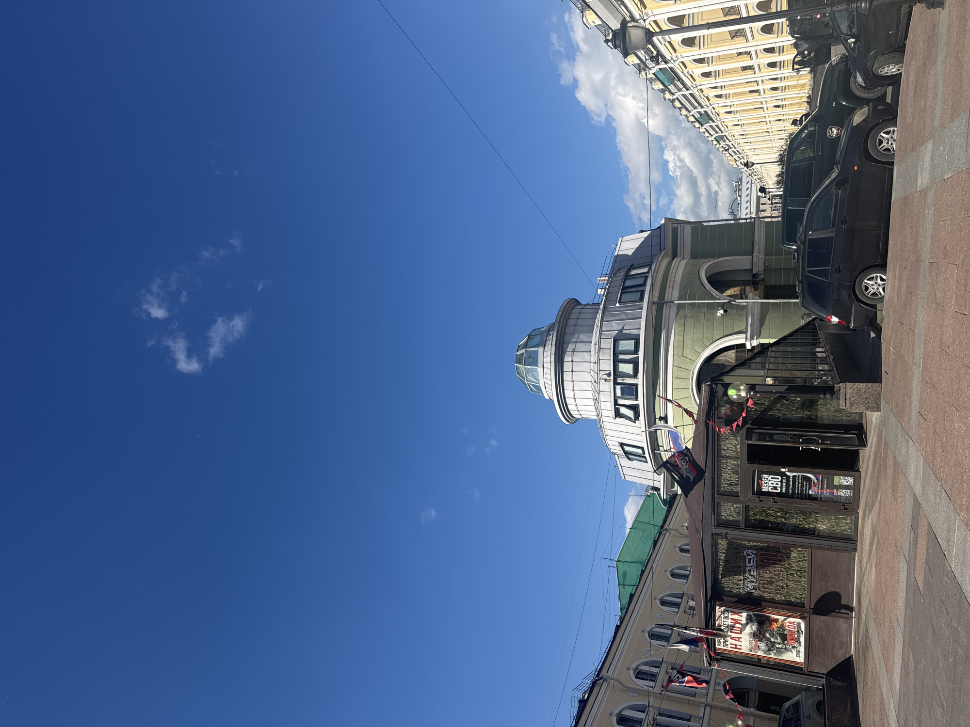 The image shows a street scene in Saint Petersburg, Russia, under a bright blue sky with scattered white clouds. In the foreground, there is a building with a glass-enclosed entrance, featuring signage indicating it is a museum labeled "МУЗЕЙ СВО". Several flags are displayed outside the building. Adjacent to this building is a green, domed structure with multiple windows. To the left and right, there are multi-story buildings with classical architectural elements. Parked cars are visible along the street. The ground is paved with stone.
