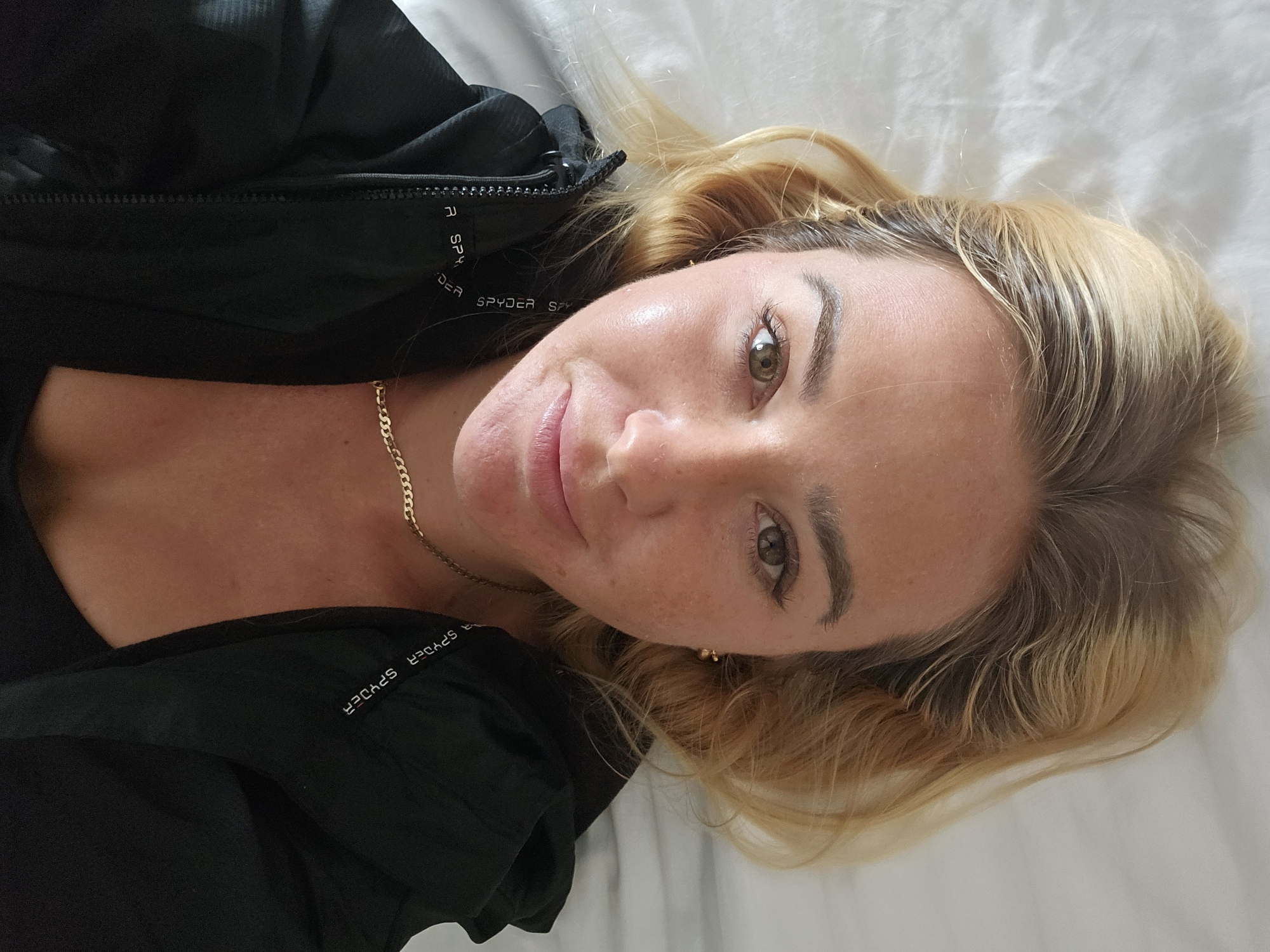 A close-up, overhead shot shows a woman lying on her back on a white textured surface. She has blonde hair, fair skin with some freckles and redness, and is wearing a black jacket with "SPYDER" written in white letters down the side. A gold chain necklace is visible around her neck. Her eyes are open and looking at the camera, and her lips are slightly parted. The background is a white, rumpled surface, likely bedding. The scene appears to be indoors, with no discernible city or environmental features. There is no indication of Da Lat, Vietnam in this image.