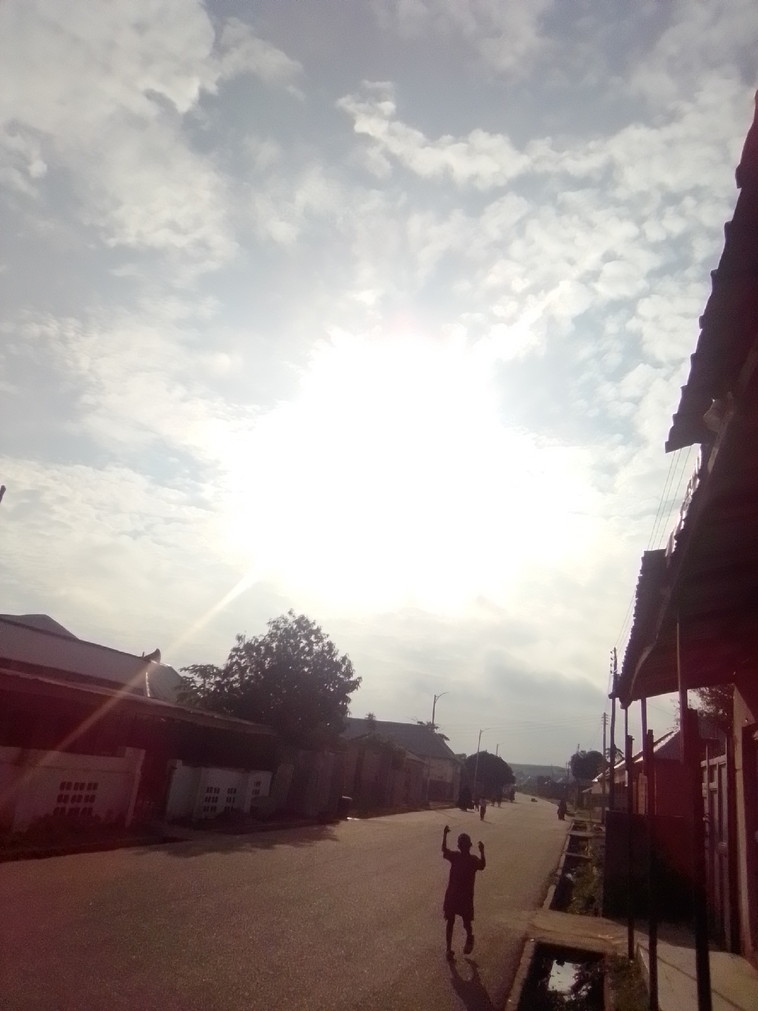 The image depicts a street scene in Liji Hill, Nigeria, under a bright, cloudy sky with the sun partially obscured. On the left side of the paved road, several buildings with corrugated metal roofs are visible, some featuring white tiled facades with dark rectangular patterns. A lone person, appearing to be a child, stands in the middle of the road with their arms raised, silhouetted against the bright sky. The road stretches into the distance, lined with more buildings and vegetation. On the right side of the road, more buildings with dark roofs and visible roof supports are present, with power lines strung between poles. A drainage channel runs alongside the road on the right. The overall impression is of a residential or commercial street in a Nigerian town, with a sense of activity suggested by the lone figure in the road.