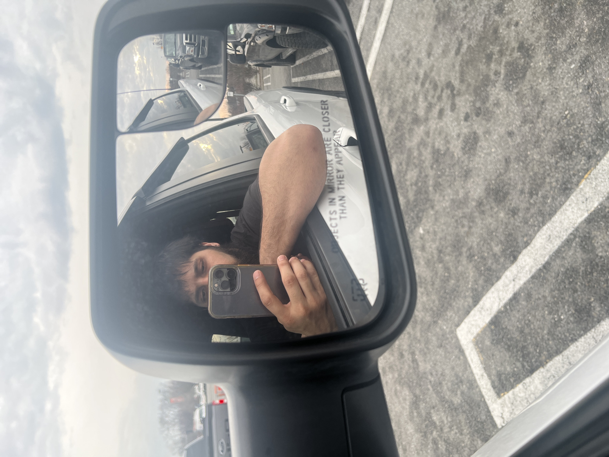The image shows a reflection in a vehicle's side mirror. The reflection captures a person holding a smartphone, presumably taking a selfie. The person has dark hair and is wearing a dark shirt. The mirror also reflects the exterior of the vehicle, including the side of the car, and the surrounding parking lot. The text "OBJECTS IN MIRROR ARE CLOSER THAN THEY APPEAR" is visible on the mirror's surface. Other vehicles are visible in the background of the mirror's reflection, suggesting a busy parking area. The location is indicated as Orange Lake, United States, based on provided data.