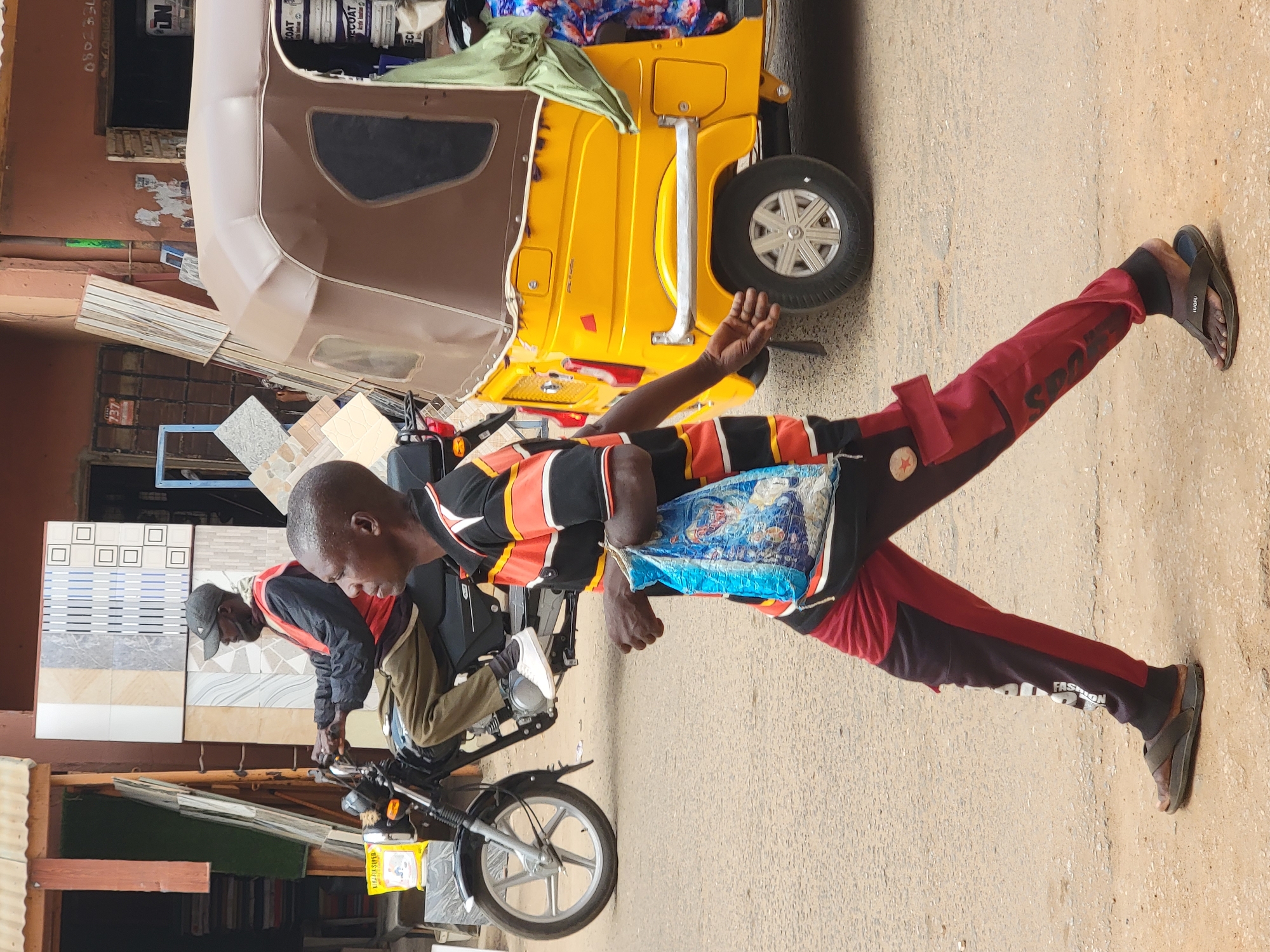 Location: Maiburiji, Nigeria.
Scene: A man wearing a striped black, orange and white shirt, red and black pants, and sandals, is walking on a paved street. He has a blue plastic bag slung over his shoulder. In the background, there is a yellow auto rickshaw, a motorcycle with two men working on it amidst stacks of tiles and other goods, and buildings.
Subjects: Man walking, two men working on motorcycle.
Objects: Auto rickshaw, motorcycle, tiles, plastic bag, sandals, clothes.
Actions: Walking, working.
Interactions: Men working on motorcycle.
Environment: Paved street, buildings, possibly a shop.