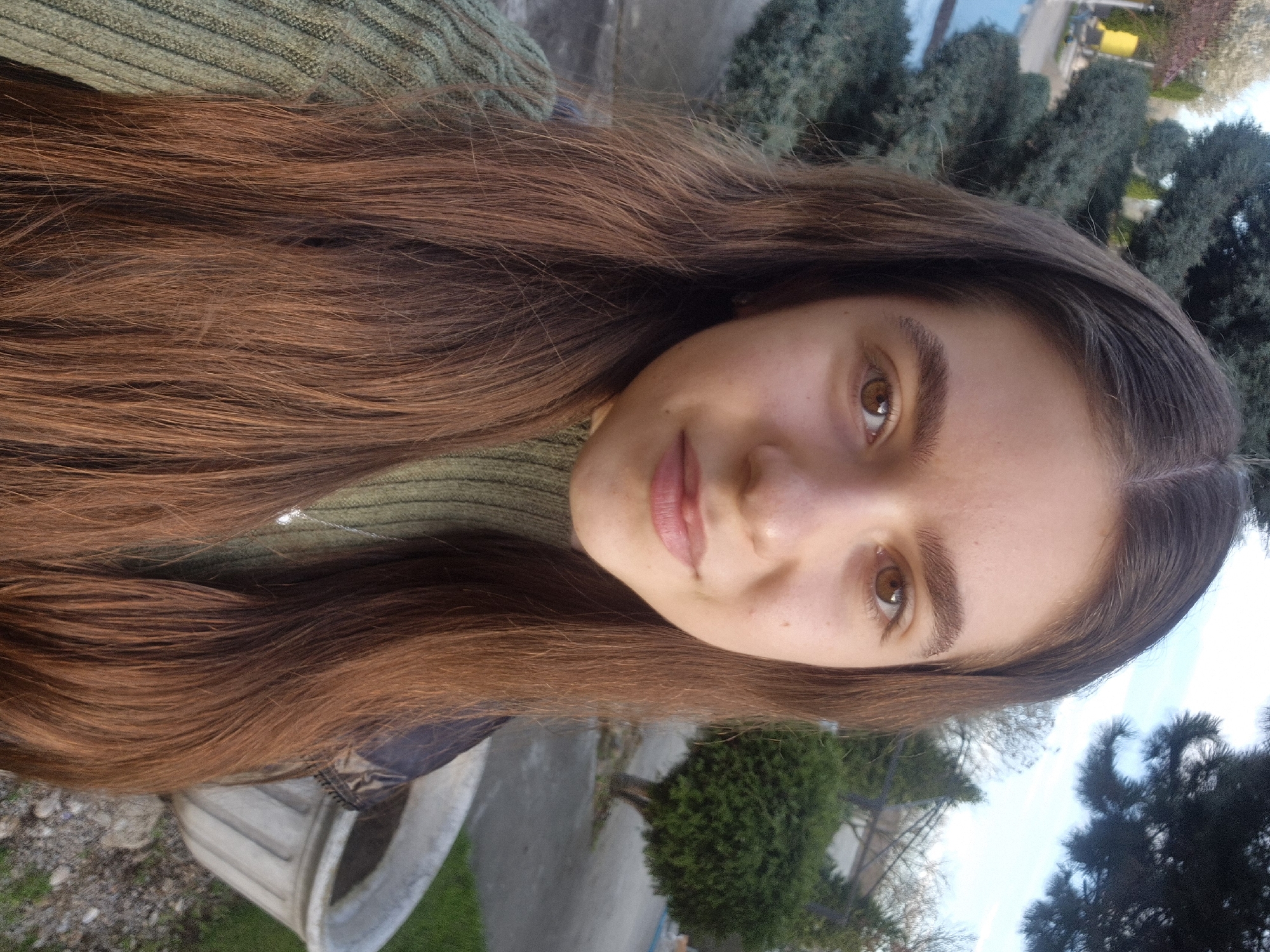 A portrait of a fair-skinned person with long brown hair and brown eyes, wearing a green ribbed sweater. The background includes evergreen trees, a light blue sky, and a glimpse of buildings and a yellow vehicle. The image appears to be taken outdoors, possibly in a park or garden area. While the city of Pavlikeni, Bulgaria is noted, there are no readily identifiable landmarks within the image to confirm this.