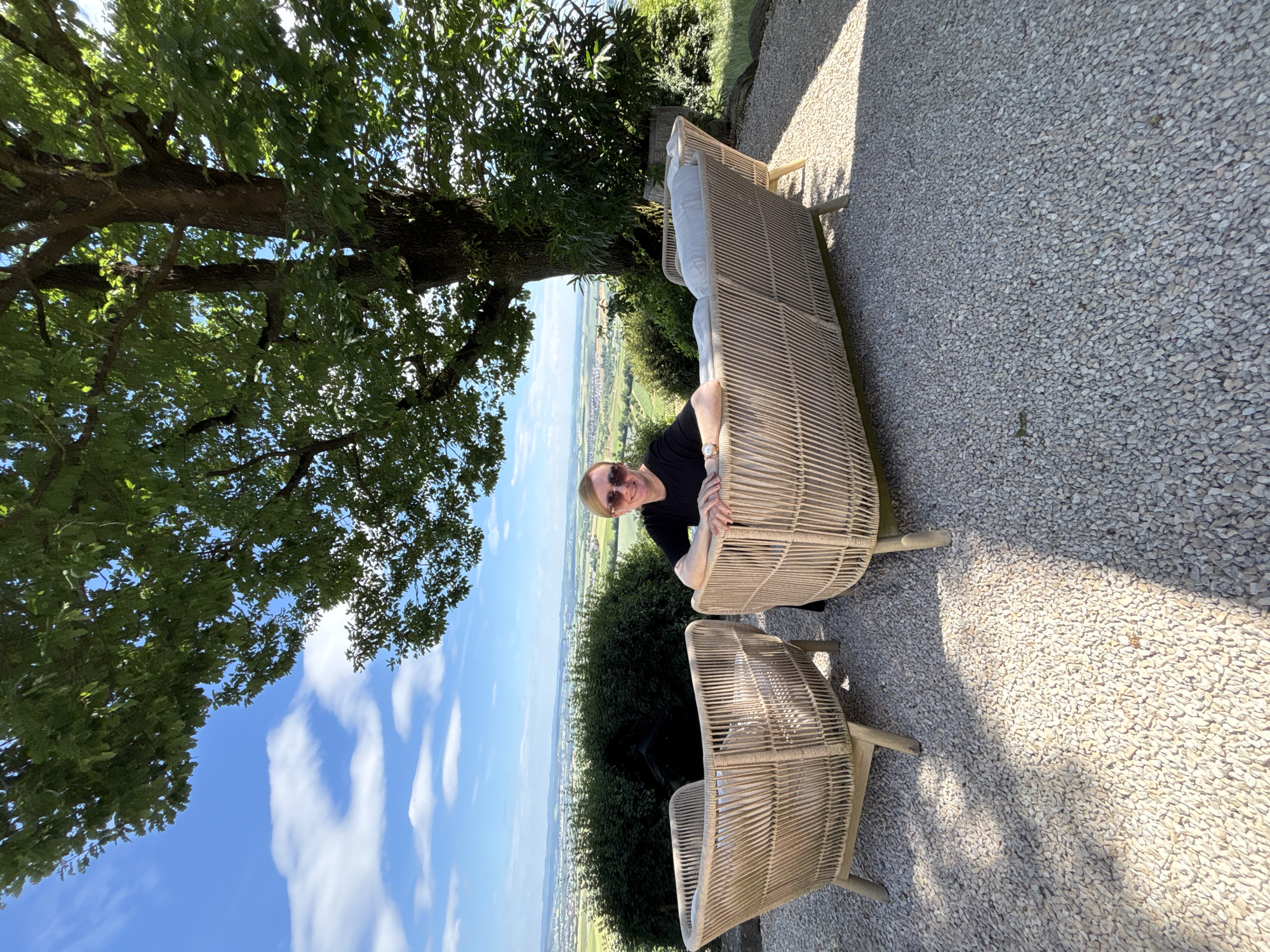 A person is sitting on a woven sofa outdoors. They are wearing sunglasses and a black top, and appear to be smiling. The sofa is light brown and has white cushions. Next to the sofa is a matching woven chair. The ground is covered in light-colored gravel. Behind the furniture, there is a lush green landscape with a tree providing shade. The sky is blue with some white clouds. The overall scene suggests a relaxed outdoor setting on a sunny day. The image is oriented vertically, with the landscape appearing at the top.
