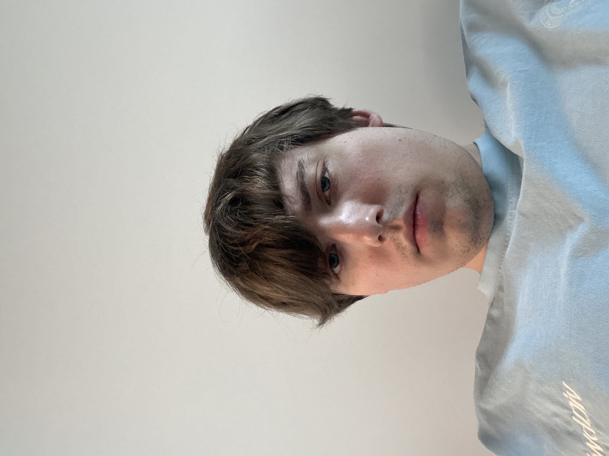 The image shows a close-up, low-angle shot of a person's face and upper body. The person has medium brown hair styled with bangs that fall over their forehead and blue eyes. They are wearing a light blue t-shirt. The background is a plain, light-colored wall. No specific location in the city of Dnipro, Ukraine can be determined from the image.