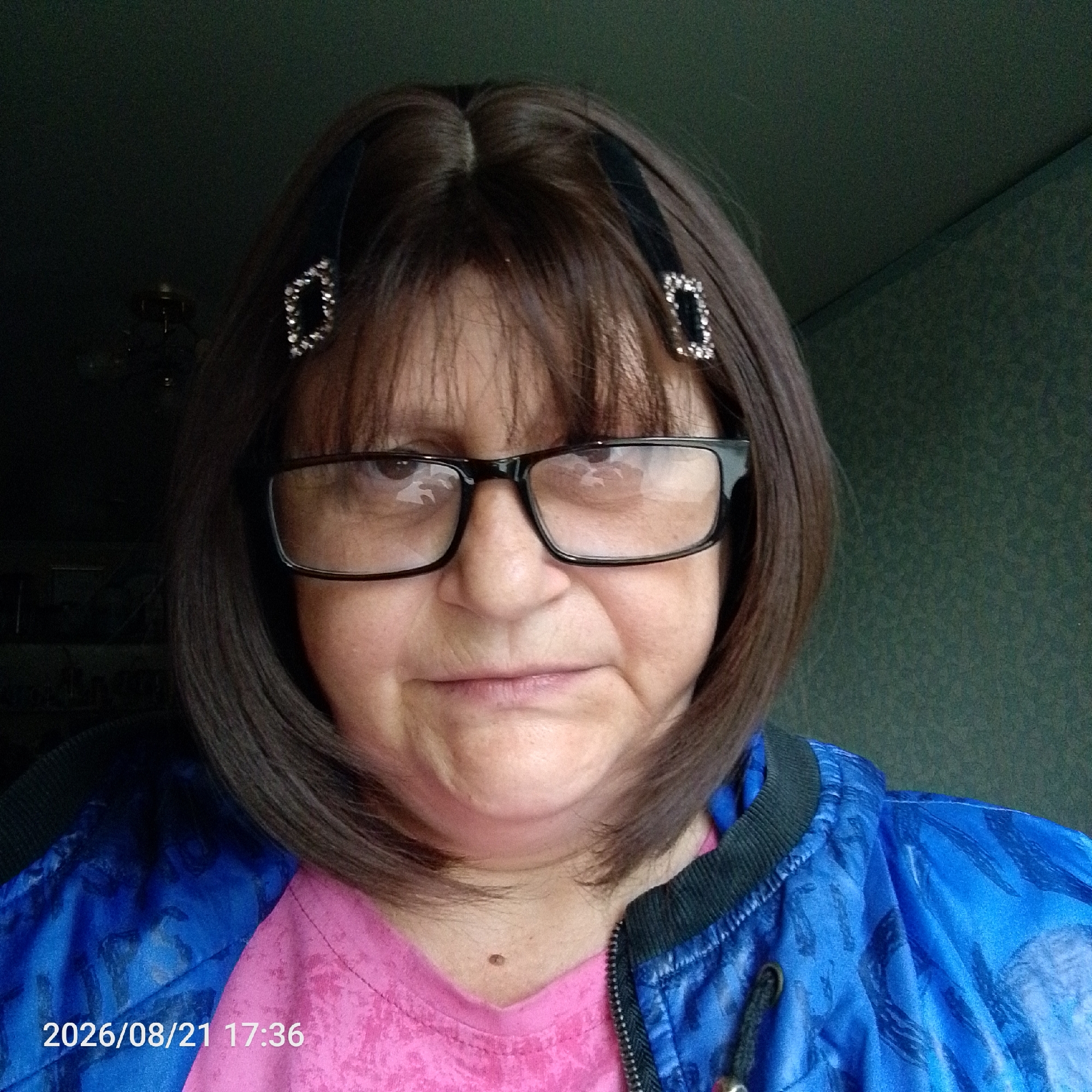 A medium shot portrait shows a woman with brown hair and glasses. She is wearing a pink shirt and a blue jacket with a patterned design. The woman has her hair pulled back on the sides with black headbands adorned with rhinestones. A date and timestamp, "2026/08/21 17:36", are visible in the lower left corner of the image. The background is a textured, dark green wall.