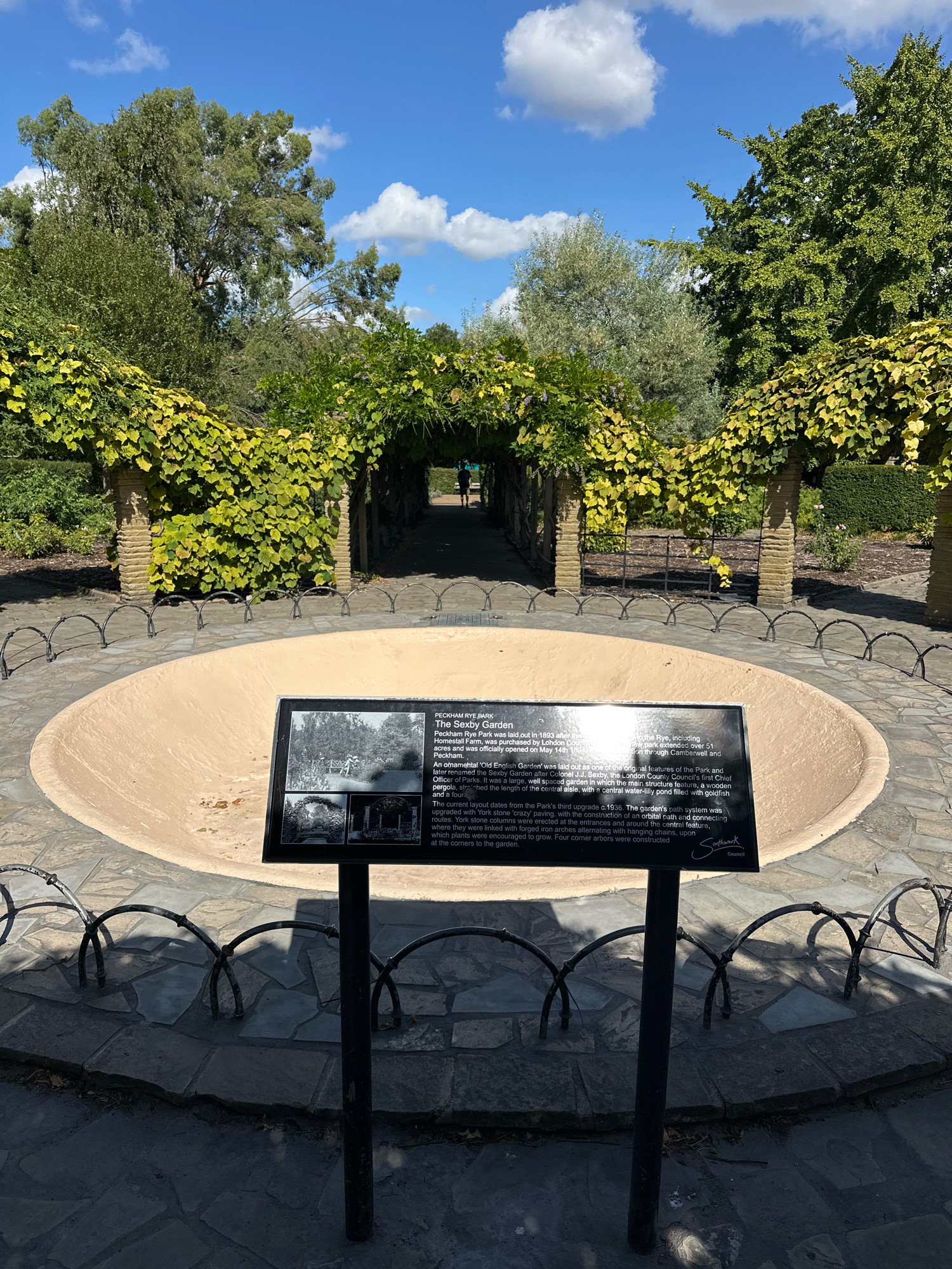 The image shows an outdoor scene in Peckham Rye Park, London, United Kingdom. In the foreground, there is a large, empty, circular basin with a beige-colored interior, bordered by a stone pathway and low, decorative black metal fencing. A signpost with a plaque is positioned in front of the basin. The plaque displays historical information and images related to "The Sexby Garden" at Peckham Rye Park. Behind the basin, a green pergola-covered walkway extends into the distance, with a person walking away from the camera in the background. The pergola is adorned with lush green and yellow foliage. Tall trees with dense green canopies are visible on either side of the walkway, and the sky above is blue with scattered white clouds. The environment appears to be a well-maintained public park on a sunny day.