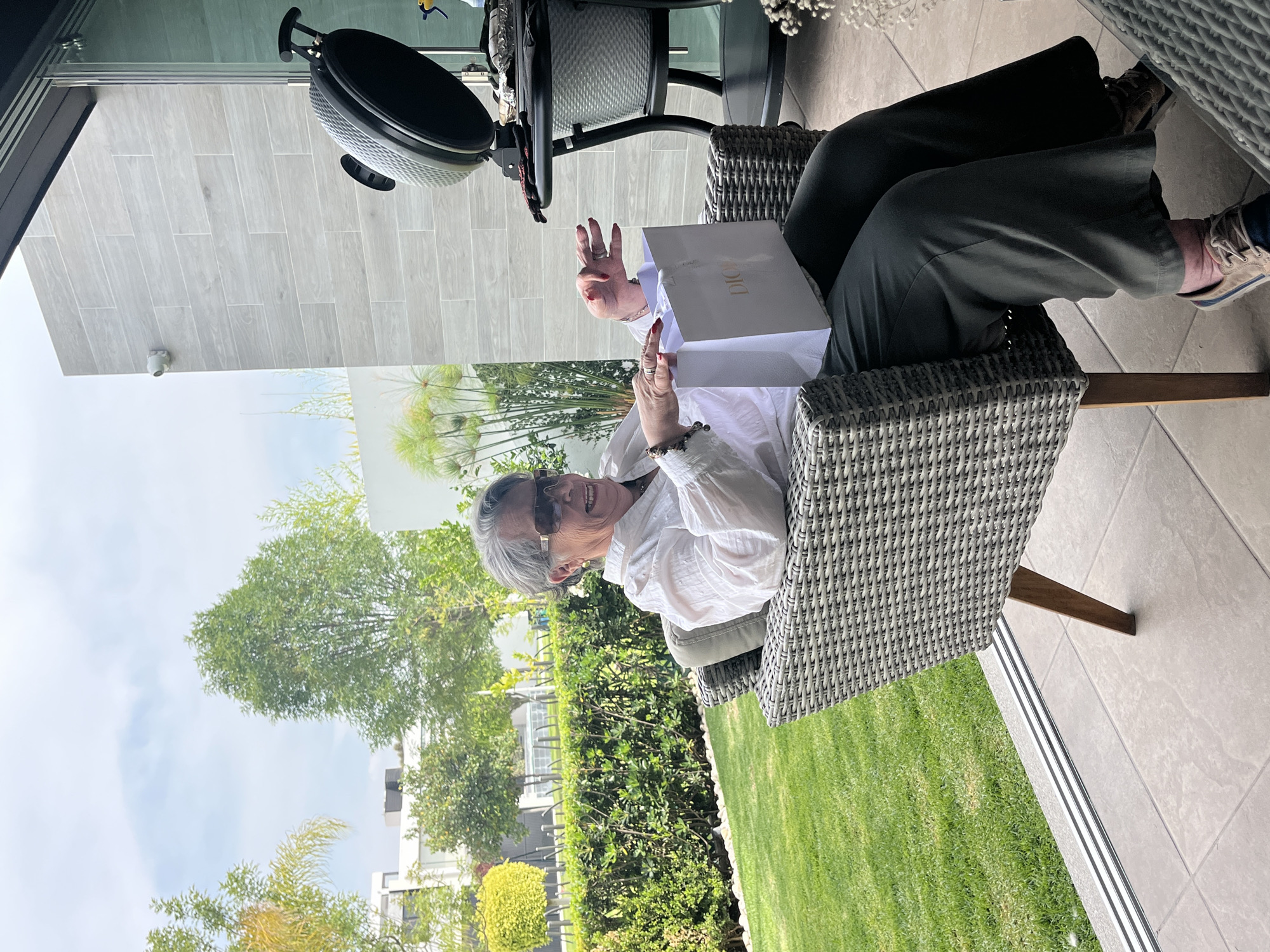 An elderly woman with short gray hair and sunglasses sits in a wicker chair outside, holding a white Dior shopping bag. She wears a white blouse and olive green pants. The background features a green lawn, hedges, and trees, suggesting a well-maintained garden or patio. The scene includes outdoor furniture and a glimpse of buildings in the distance. The location is most likely in San Bernardino Tlaxcalancingo, Mexico.