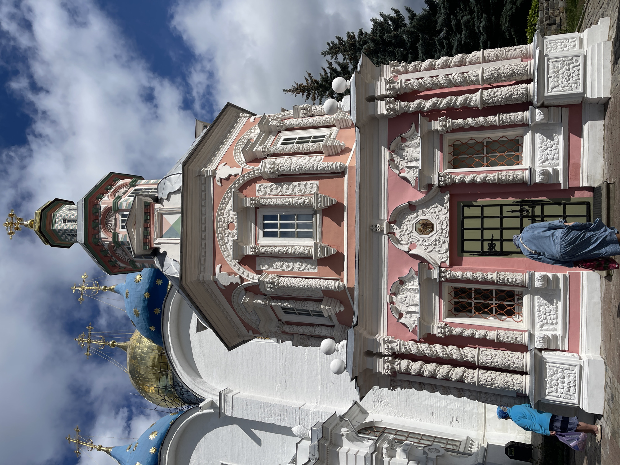 This is a low-angle, full shot of the Church of the Nativity of the Virgin, a part of the Trinity Lavra of St. Sergius complex in Sergiev Posad, Russia. The church features a distinctive pink facade with white ornate stucco decorations, including twisted columns and angelic figures. Several windows with ornate iron grates are visible. A green metal door is present at the entrance. Two individuals are seen from behind. One person, wearing a blue jacket and headscarf, stands to the left, holding shopping bags. The second person, in a blue denim jacket and patterned skirt, is walking towards the entrance. In the background, to the left, are portions of a larger white church with bright blue domes adorned with gold stars, and a golden dome. A tall evergreen tree stands to the right. The sky is partly cloudy with patches of blue.