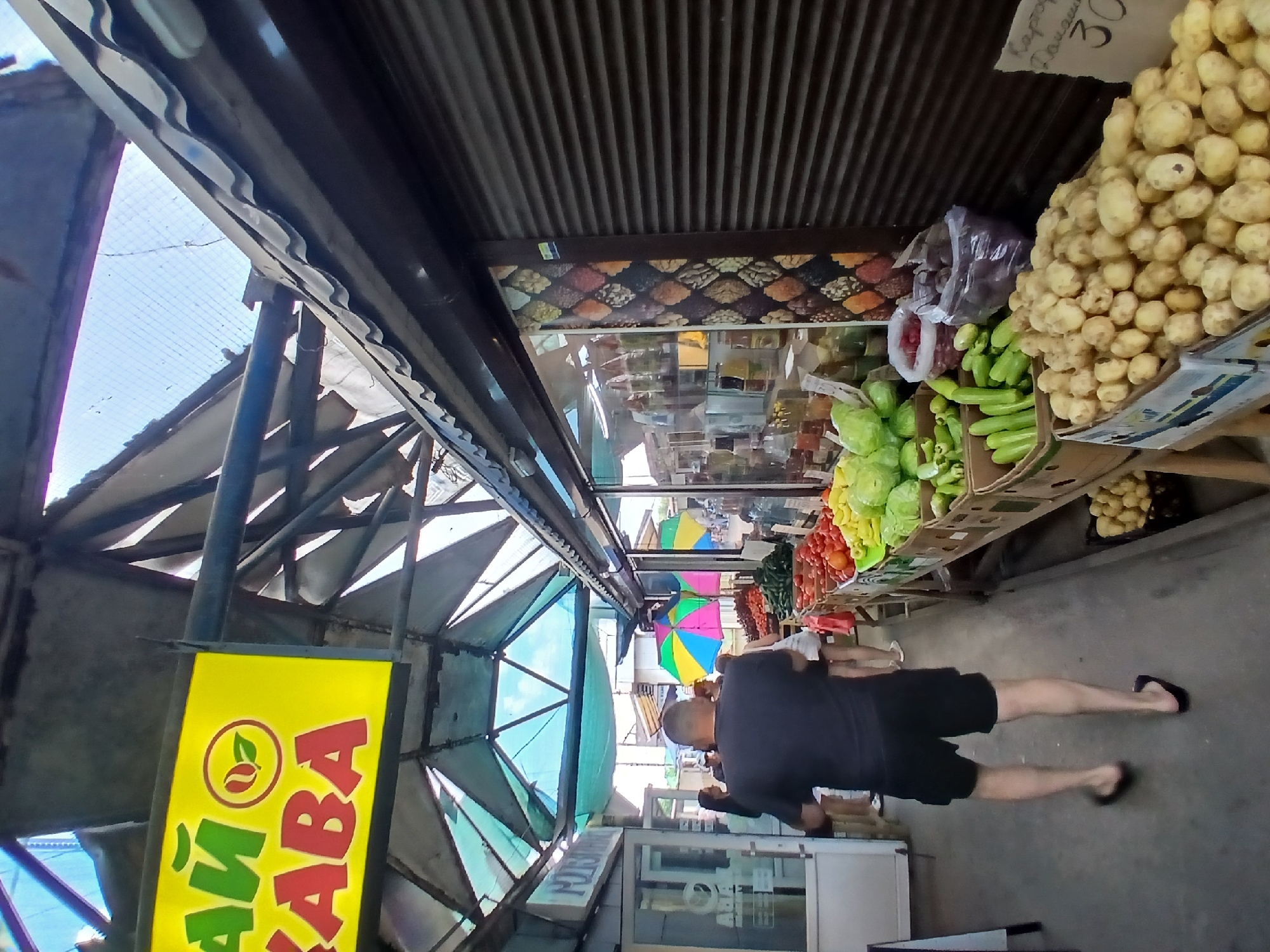 The image depicts a market stall in Dnipro, Ukraine, likely an outdoor or semi-outdoor market given the visible roofing structure. The foreground features a man in a black t-shirt and shorts walking away from the camera, wearing flip-flops. To his right, a market stall is laden with fresh produce, including boxes of potatoes in the immediate foreground, followed by rows of cucumbers, green peppers, tomatoes, and cabbage. Further down the stall, there are visible signs and umbrellas, suggesting a lively market atmosphere. The stall on the right has a rolled-down metal shutter and a patterned decorative panel. The background shows other market stalls and shoppers, with a bright sky visible through a translucent, geometric roofing system. A yellow sign with green and red lettering, which appears to read "ЧАЙ КАВА" (Tea Coffee), is prominently displayed overhead. The overall scene is one of a typical bustling marketplace.