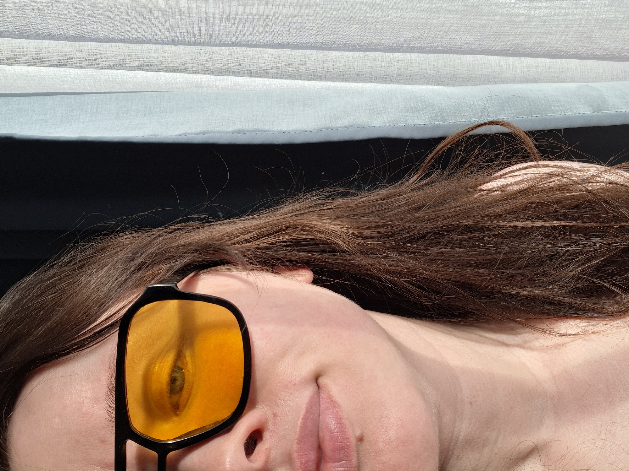 A close-up, portrait-style photograph shows a person lying down. The person has long, brown hair and is wearing yellow-tinted sunglasses. The eye reflected in the sunglasses is visible. Above the person is a gauzy, layered curtain in white and light blue, with a dark space visible behind it. The location and city are unknown.