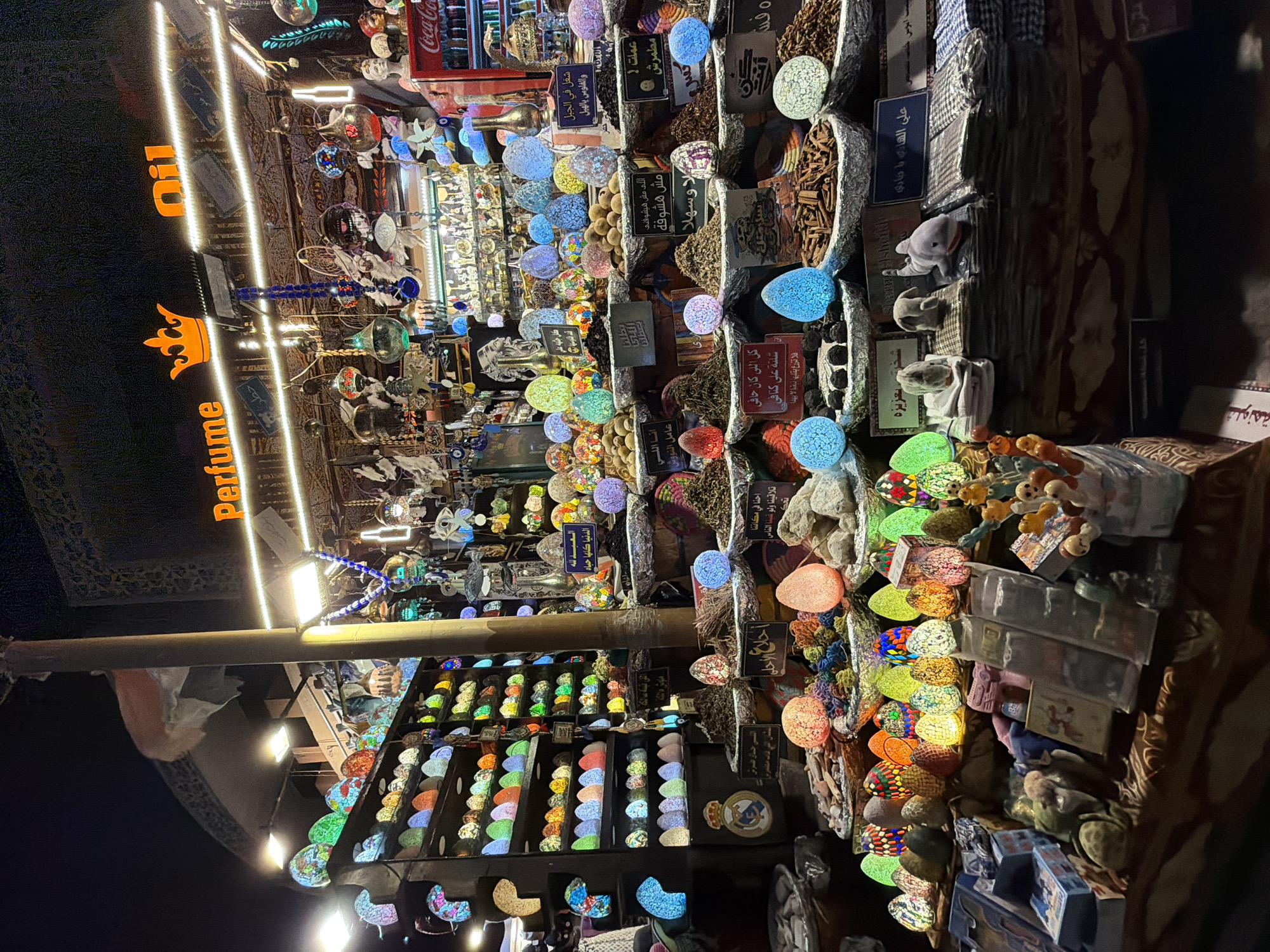 This is a night-time, eye-level, medium shot of a stall in Sharm El Sheikh, Egypt, that appears to sell perfumes, oils, and various decorative items. The stall is brightly lit with numerous colorful, egg-shaped lamps, globes, and hanging ornaments. In the background, a sign reads "Perfume Oil" with a crown emblem. Shelves behind the display are filled with more of these illuminated decorative objects, predominantly in shades of blue, green, yellow, orange, and pink.
In the foreground, a wide variety of items are displayed, including piles of spices or dried herbs in shallow, silver-rimmed baskets, and several small, plush animal toys, including a shark and a donkey, placed on folded fabric. Small signs with Arabic text are visible in front of the displays of spices. To the right of the stall, a red Coca-Cola refrigerator is partially visible. The overall atmosphere is vibrant and busy, characteristic of a market or bazaar setting at night.