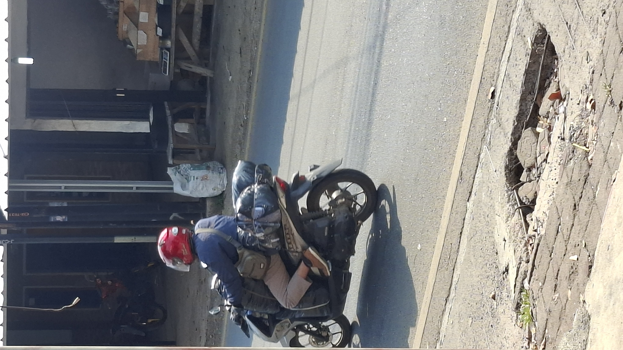 In the city of Cilaku, Indonesia, a person wearing a red helmet rides a motorcycle loaded with luggage. The rider is dressed in a blue long-sleeved shirt and light brown pants, with a tan shoulder bag visible. The motorcycle is silver and white, with several black bags and bundles secured to the rear. The motorcycle is traveling on a paved road, which appears to be in moderate condition, with some debris and a pothole visible in the foreground on the right side of the image. In the background, there are buildings with corrugated metal roofs and open storefronts, suggesting a commercial area. A large white bag of what appears to be rice or grain is placed near one of the buildings.