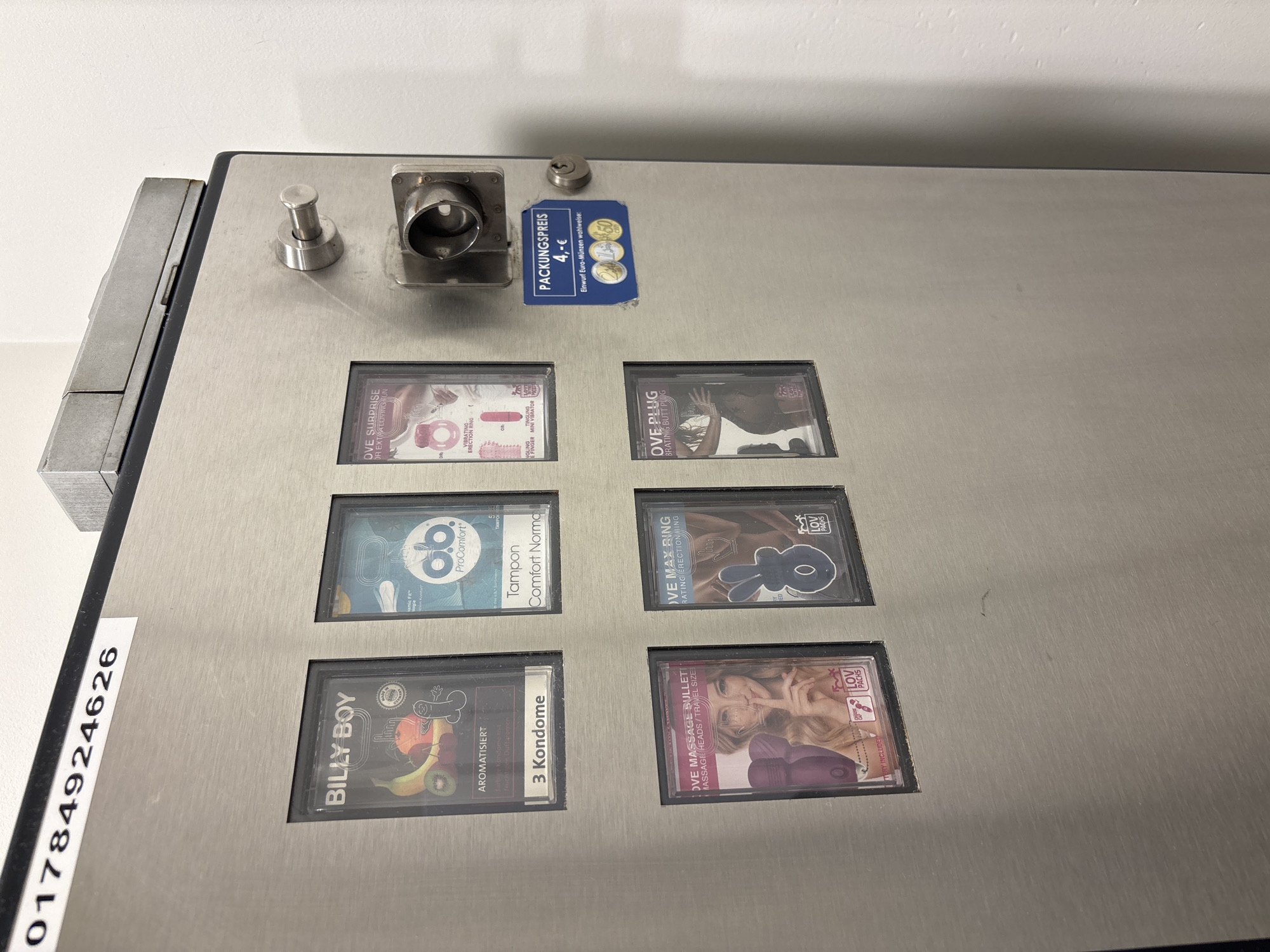 The image shows a vending machine located in Schönefeld, Germany, likely in a public restroom or establishment. The machine is made of brushed stainless steel and displays various adult novelty items and condoms.
The top row of the machine features:
- A product labeled "BILLY BOY AROMATISIERT 3 Konfome" which appears to be flavored condoms.
- A product labeled "ob ProComfort Tampon Comfort Norma."
- A product labeled "OVE SURPRISE OR EXTRA LOVING FUN VIBRATING ERECTION RING."
The bottom row of the machine features:
- A product labeled "OVE MASSAGE BULLET MASSAGE HEADS / TRAVEL SIZE."
- A product labeled "OVE MAX RING VIBRATING ERECTION RING."
- A product labeled "OVE PLUG BRATING BUTT PLUG."
To the right of the product displays, there is a coin slot and a keyhole. A sticker below the coin slot reads "PACKUNGSPREIS 4,- €," indicating the price for items and that payment can be made with Euro coins, with images of a 1 Euro, 2 Euro, and 50 cent coin shown.
A serial number or identification code, "01784924626", is affixed to the top left of the machine. The environment appears to be indoors, with a white wall visible to the right of the machine. The overall impression is a functional vending machine dispensing personal items.