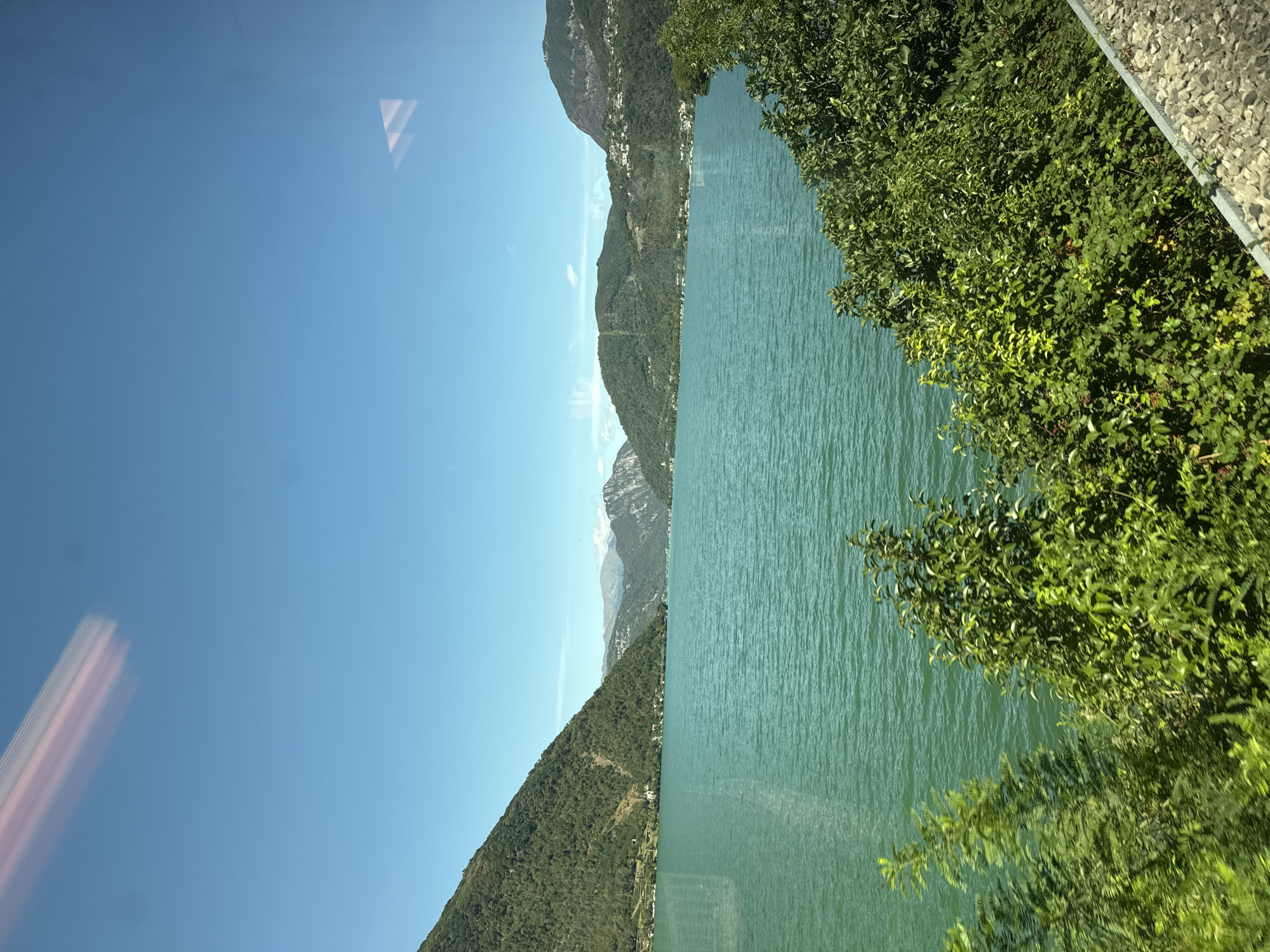 The image captures a scenic view of Lake Lugano, likely from a vantage point in or near Mendrisio, Switzerland. The water of the lake is a vibrant turquoise, rippling gently under a clear, bright blue sky. Lush green hills and mountains, densely covered with trees, surround the lake, forming a dramatic backdrop. In the distance, several mountain ranges are visible, some appearing hazy with their peaks touching the sky. Along the slopes of the closer hills, scattered buildings and residences are discernible, suggesting the presence of human settlements. In the foreground, dense green foliage, likely trees and bushes, partially obscures the view, with some appearing to be on a slope leading down towards the water. A light-colored, textured surface, possibly a path or embankment made of gravel and stone, is visible at the bottom right of the image. The overall impression is one of a serene, natural landscape bathed in sunlight. There are no people or specific actions depicted in the image.