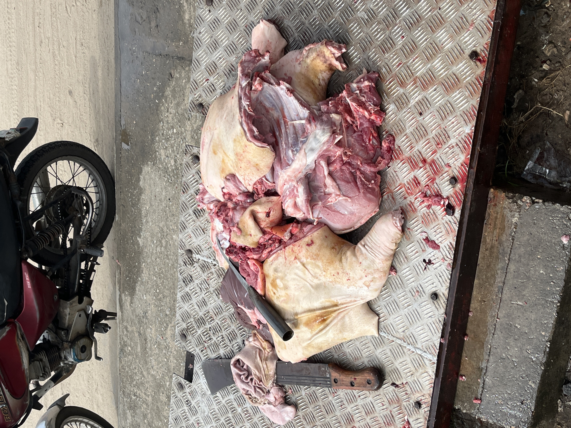 The image shows butchered pig parts on a metal surface, likely the bed of a small trailer or truck. Visible are various cuts of pork, including portions of the skin, meat, and organs. Two butchering tools, a knife and a cleaver, rest on the meat. Beside the table is a motorbike, red in color. The scene is outdoors, in a setting that appears to be urban. The specific location is Badagry, Nigeria.