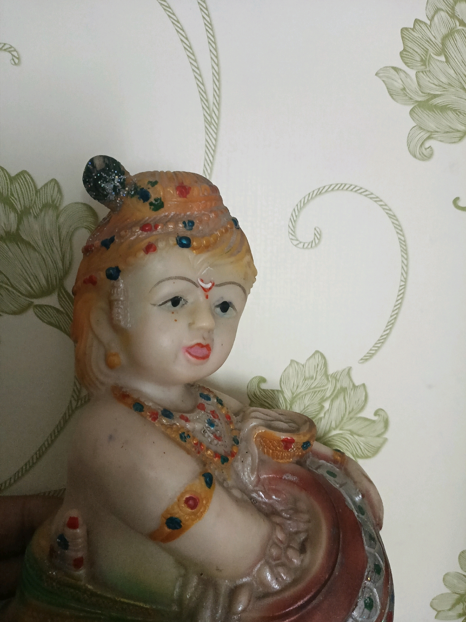 A statue of a child deity, likely Krishna, is displayed against a white wallpaper with a green floral pattern and rope-like design. The statue is made of a pale material and is adorned with colorful paint and glitter, depicting jewelry and clothing. The deity has a small bun of hair on its head, painted orange with some glitter. The eyes are dark, and there is a red dot on the forehead. The deity is holding a brown pot with a red rim, and its arms and legs are visible. The background wallpaper features stylized green leaves and swirling lines. The statue appears to be a decorative item, possibly placed on a shelf or table. No specific location in Manpur, India is indicated by the visual information in the image.