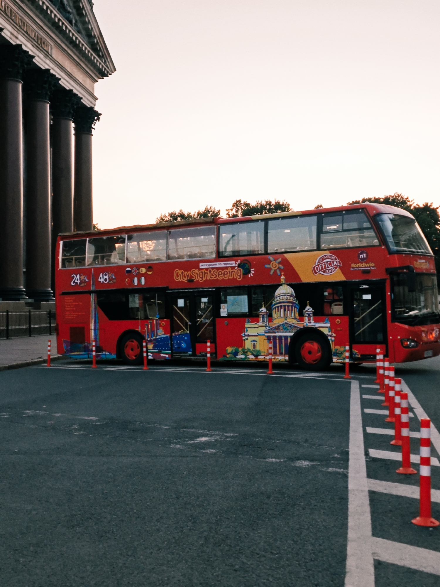 This image captures a red double-decker "City Sightseeing" tour bus parked on an asphalt road in Saint Petersburg, Russia. The prominent classical building on the left is Saint Isaac's Cathedral, characterized by its massive, dark columns and a visible portion of its entablature and pediment.
The red double-decker bus features "City Sightseeing" branding in yellow, alongside "OFFICIAL TOUR" and "Worldwide Sightseeing Tours" logos. The side of the bus displays colorful illustrations of Saint Petersburg landmarks, including a detailed graphic of Saint Isaac's Cathedral itself. Numbers "24" and "48" are visible, accompanied by a row of national flags indicating multilingual services, including Russian, British, German, Italian, Chinese, and Japanese. The upper deck of the bus is visible, with reflective windows.
The road is dark asphalt, marked with white painted lines defining a designated area. A series of orange and white striped flexible bollards are uniformly arranged along one of the white lines, creating a barrier or guiding path. Behind the bus, a line of dark green trees is visible against a clear, light-toned sky, suggesting either dawn or dusk lighting conditions. No individuals are visible in the scene.