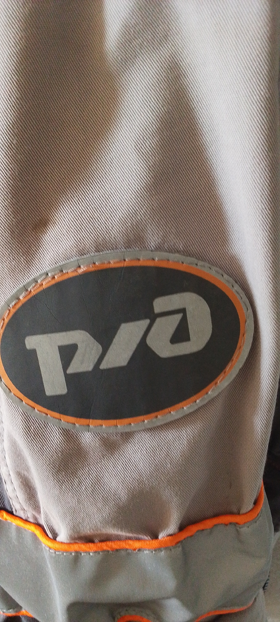 A close-up image shows a patch on a grey garment. The patch is oval-shaped and bordered by orange stitching. Inside the oval, a dark grey surface features a light grey logo that appears to read "P/A". The background is a textured, light brown fabric. The image is a detail shot and does not depict any city, environment, or identifiable location in Bira, Russia. There are no people, actions, or interactions visible.