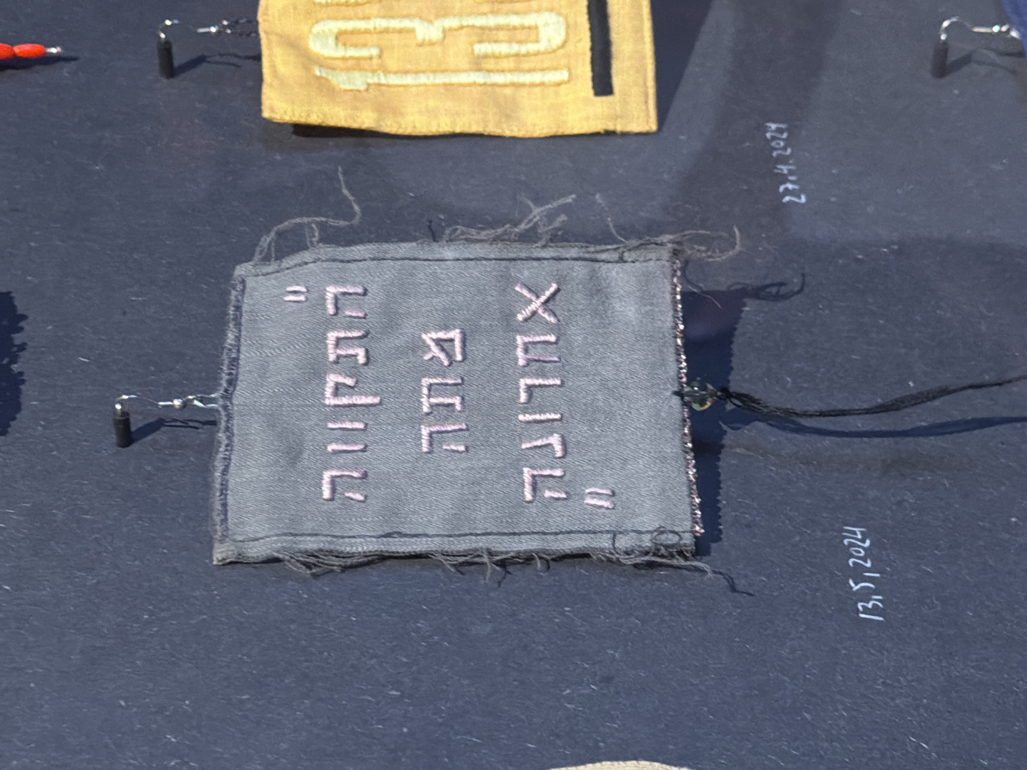 The image depicts a close-up view of a gray, fabric tag with Hebrew text stitched in pink. The text reads: "אֲדָנָי שְׂפָתַי תִּפְתָּח" which translates to "My Lord, open my lips". The tag is pinned to a dark surface, likely part of an exhibit. A separate tag displaying the number "13" in yellow is visible in the upper portion of the image. Handwritten dates "27.4.2024" and "13.5.2024" are written to the side of the tag. The setting appears to be a museum exhibit in Tel-Aviv, Israel.
