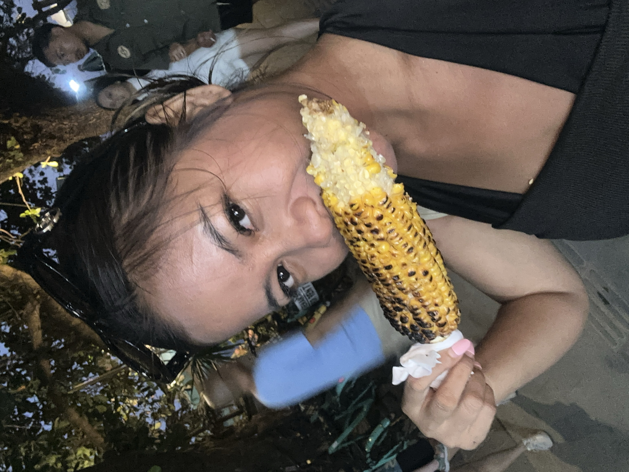 A woman with dark hair and a black top is taking a selfie as she eats a cob of grilled corn. She is holding the corn with her right hand, which is wrapped in white tissue. Her left hand is not visible. She is looking at the camera with a smile and has pink nail polish. In the background, a man in a green uniform with a badge is standing next to a woman in a white dress. Trees with leaves are visible above and behind them. The ground appears to be paved. The overall lighting suggests it is evening. The scene is likely at a street food vendor or an outdoor market in Denpasar, Indonesia.