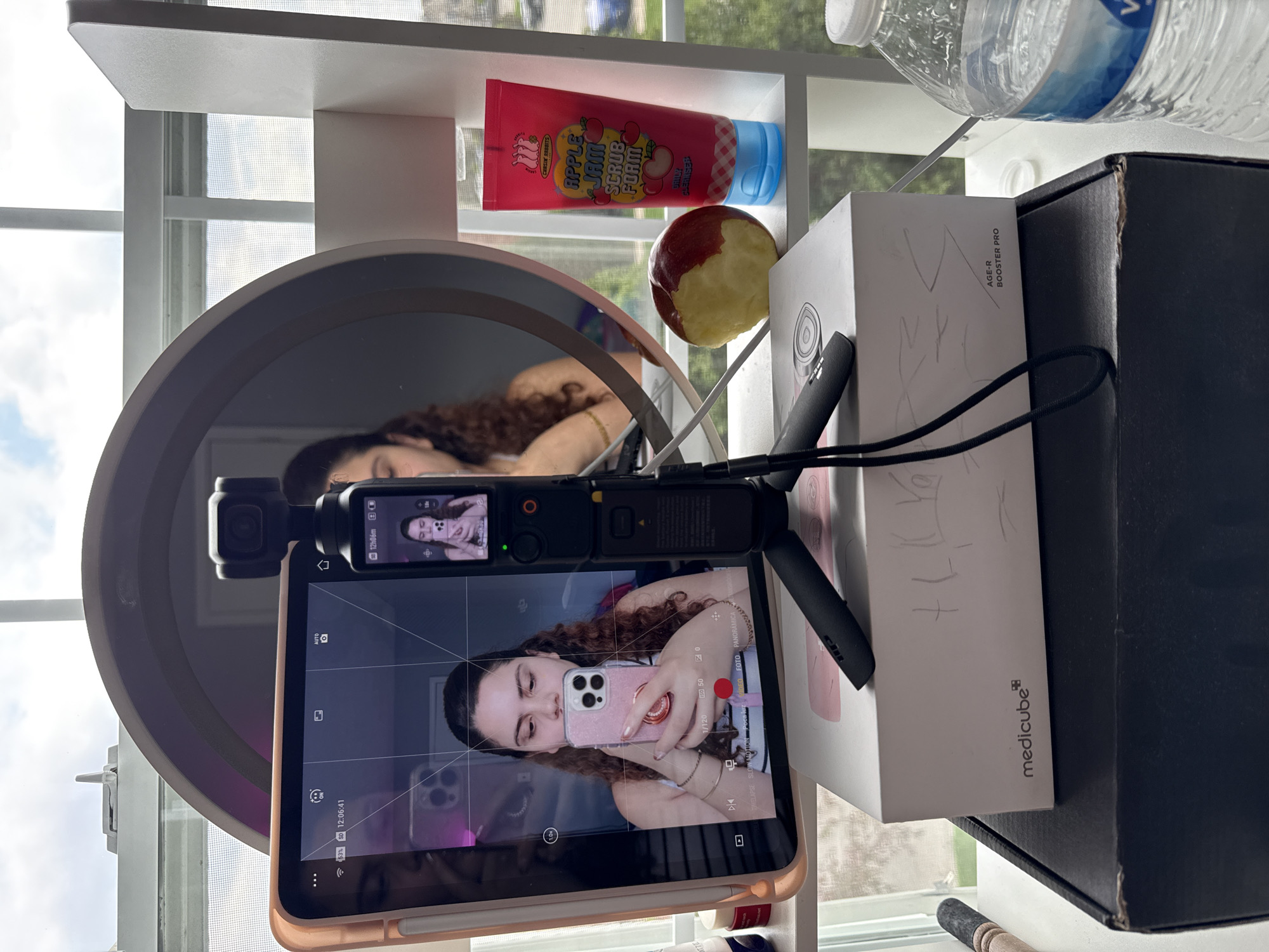 A vertically oriented close-up shot depicts an arrangement of objects on a shelf, with a focus on reflections and technology. The shelf, situated in front of a window, holds a variety of items, including a mirror reflecting a person with curly brown hair. The reflection shows the person lying down, with a gold bracelet visible on their wrist.
In the foreground, a phone or tablet displays a live view of the same person holding a pink phone, positioned as if taking a selfie. The device is connected to a black camera stabilizer on a tripod.
Behind the phone, the shelf contains a tube of red face scrub labeled "APPLE JAM SCRUB FORM," along with an apple with a bite taken out of it. To the right, there is a white box with the text "AGE-R BOOSTER PRO" and "medicube" visible, sitting next to a larger black box. A bottle of water is placed near the top right corner of the frame. The overall composition suggests a behind-the-scenes view of content creation or a personal video setup. The environment appears to be indoors, near a window with a view of the outside, but the exact location is not discernible from the image.