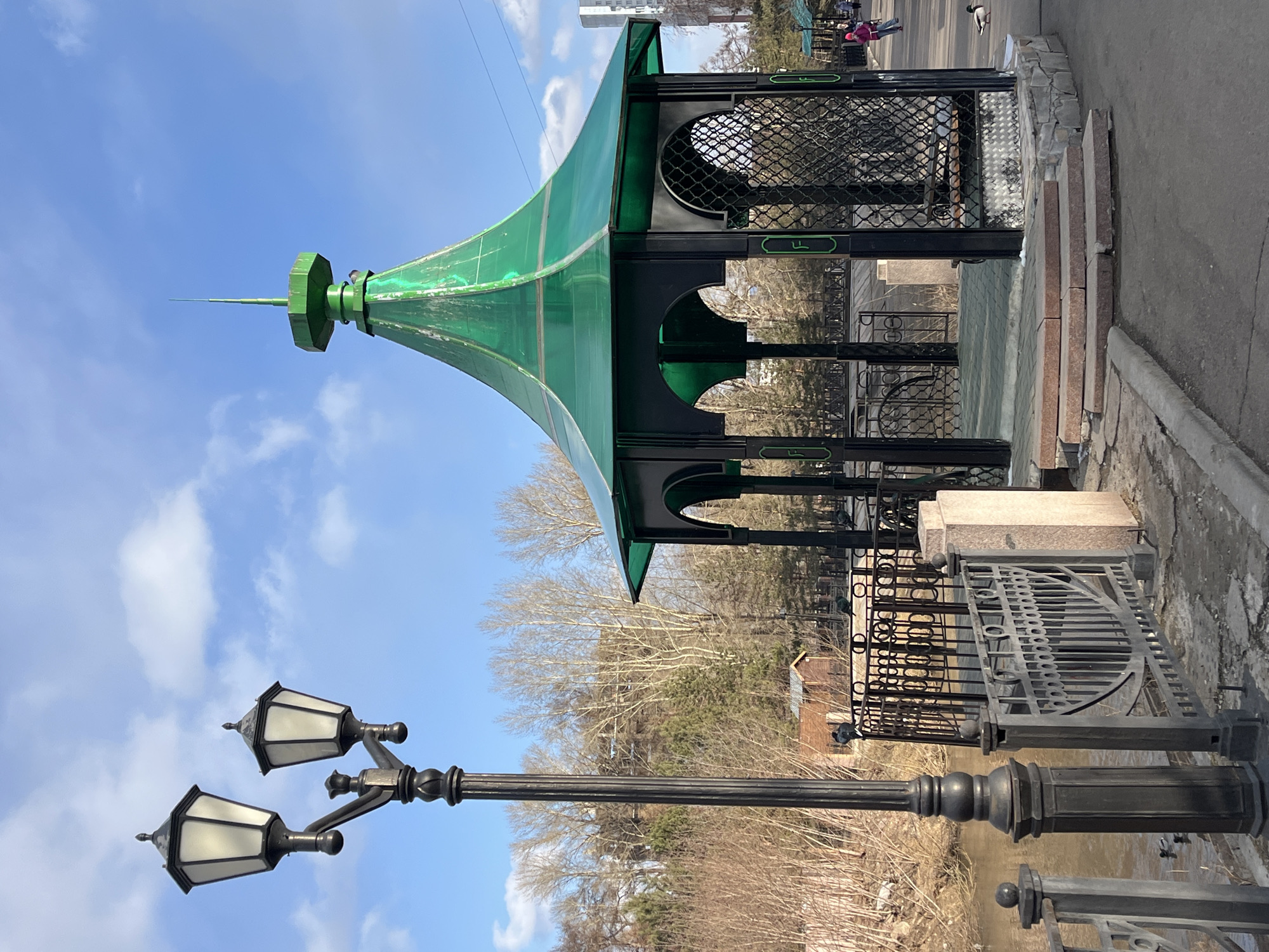 The image features a green-roofed gazebo located in the city of Krasnoyarsk, Russia. The gazebo has black metal arches and a decorative metal screen along the top. Adjacent to the gazebo is a black, dual-lamp street light with ornate detailing. The setting appears to be a park or public area with a body of water visible below the railing next to the gazebo. Some bare trees are also visible in the background. A couple of individuals walking and a duck can be seen in the upper right corner of the image. The sky is light blue with scattered clouds. The image is vertically oriented.