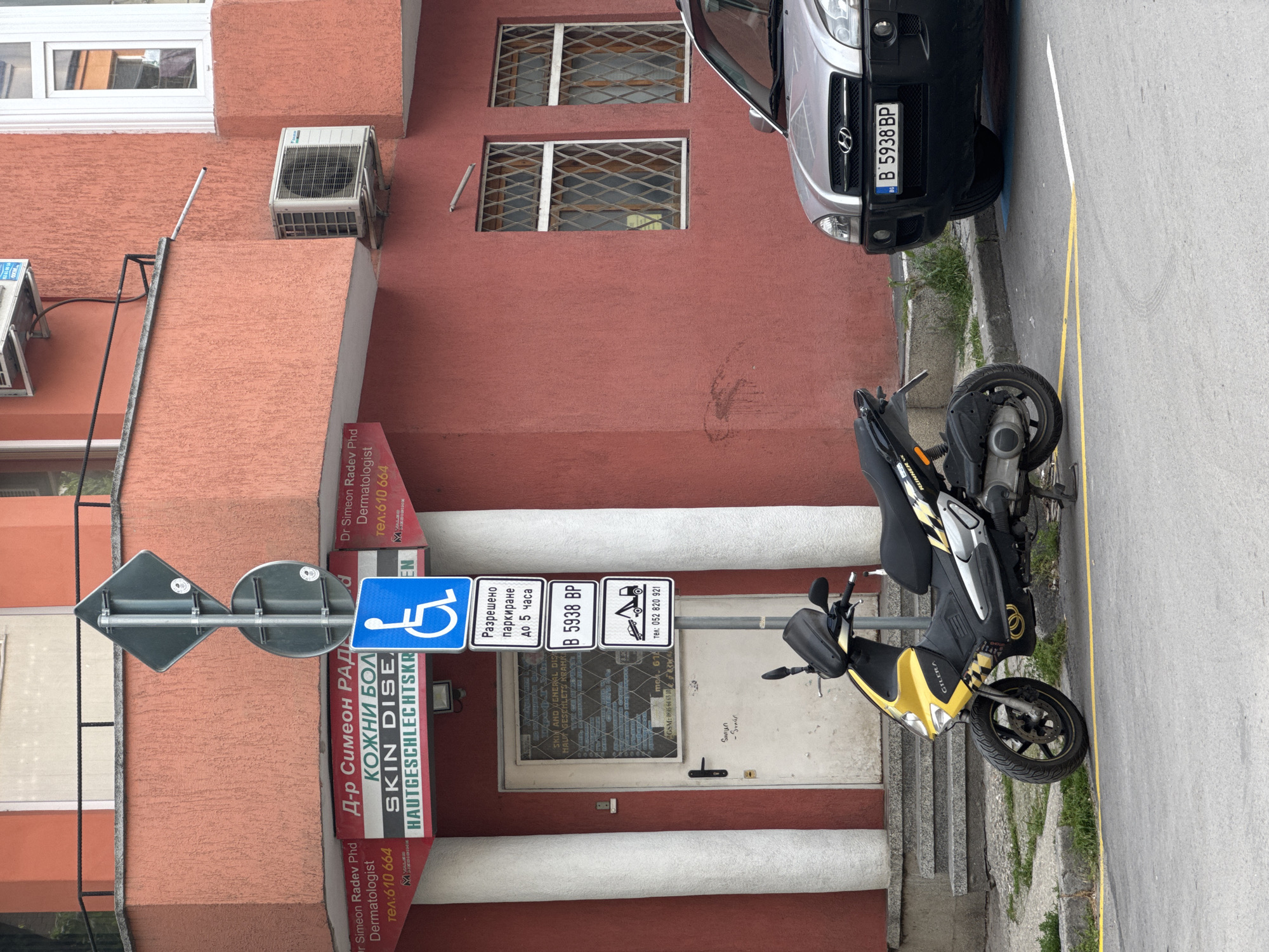 This eye-level, outdoor shot depicts a street scene in Varna, Bulgaria, during daytime under what appears to be overcast skies. The main subject is a reddish-brown stucco building, likely a commercial establishment, with a sidewalk and street in front of it.
A yellow and black Gilera Runner scooter is parked on its kickstand on the sidewalk. Partially visible on the street is the front of a silver Hyundai car, bearing the Bulgarian license plate "B 5938 BP". The street features yellow lines, possibly delineating parking spaces or a lane.
The building's facade includes multiple windows, some fitted with metal security grates, and two external air conditioning units. A white entrance door is framed by two white columns.
Several signs provide information about the location. A large vertical red and white sign on the building identifies the practice as "Д-р Симеон РАДЕВ" (Dr. Simeon Radev), a "Dermatologist" specializing in "КОЖНИ БОЛ" (Skin Diseases), "SKIN DISE", and "HAUTGESCHLECHTSKR" (Venereal Diseases in German), with a contact number "тел:610 664". Another sign on the entrance door further details "SKIN AND WENERAL DIS" and "HAUT GESCHLETS KRANKI" (likely a partial German translation), along with mobile and landline numbers: "GSM: 0885 64 65 10" and "тел: 052 820 921".
Mounted on a pole in front are multiple street signs: a blue handicap parking sign, a white sign indicating "Разрешено паркиране до 5 часа" (Allowed parking up to 5 hours), another sign displaying the license plate "B 5938 BP", and a sign featuring a tow truck icon with "тел: 052 820 921", signifying a tow-away zone. No people are present in the static scene.