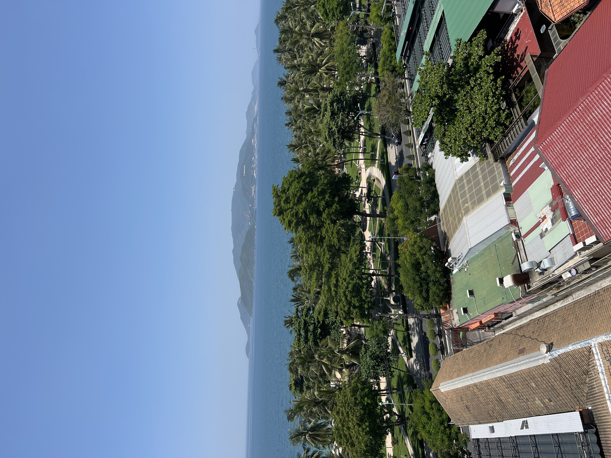The image depicts a coastal city scene with a clear blue sky above a wide expanse of calm, turquoise water. In the distance, a long, low mountain range forms the horizon. Closer to the foreground, a dense line of lush green palm trees and other foliage separates the ocean from a park area. This park features manicured lawns, pathways, and several trees. Beyond the park, a street runs parallel to the coast, with a few cars visible. The immediate foreground is dominated by the rooftops of buildings, characterized by their tiled surfaces in shades of brown, red, and green, as well as metal sheeting. Air conditioning units and other rooftop fixtures are also present. The overall impression is of a sunny day in a tropical urban environment, likely a resort area. The presence of palm trees, the ocean, and the mountainous backdrop strongly suggest a location along the coast of Vietnam, specifically Nha Trang, which is known for its beaches and coastal scenery. No people are explicitly visible in the image.