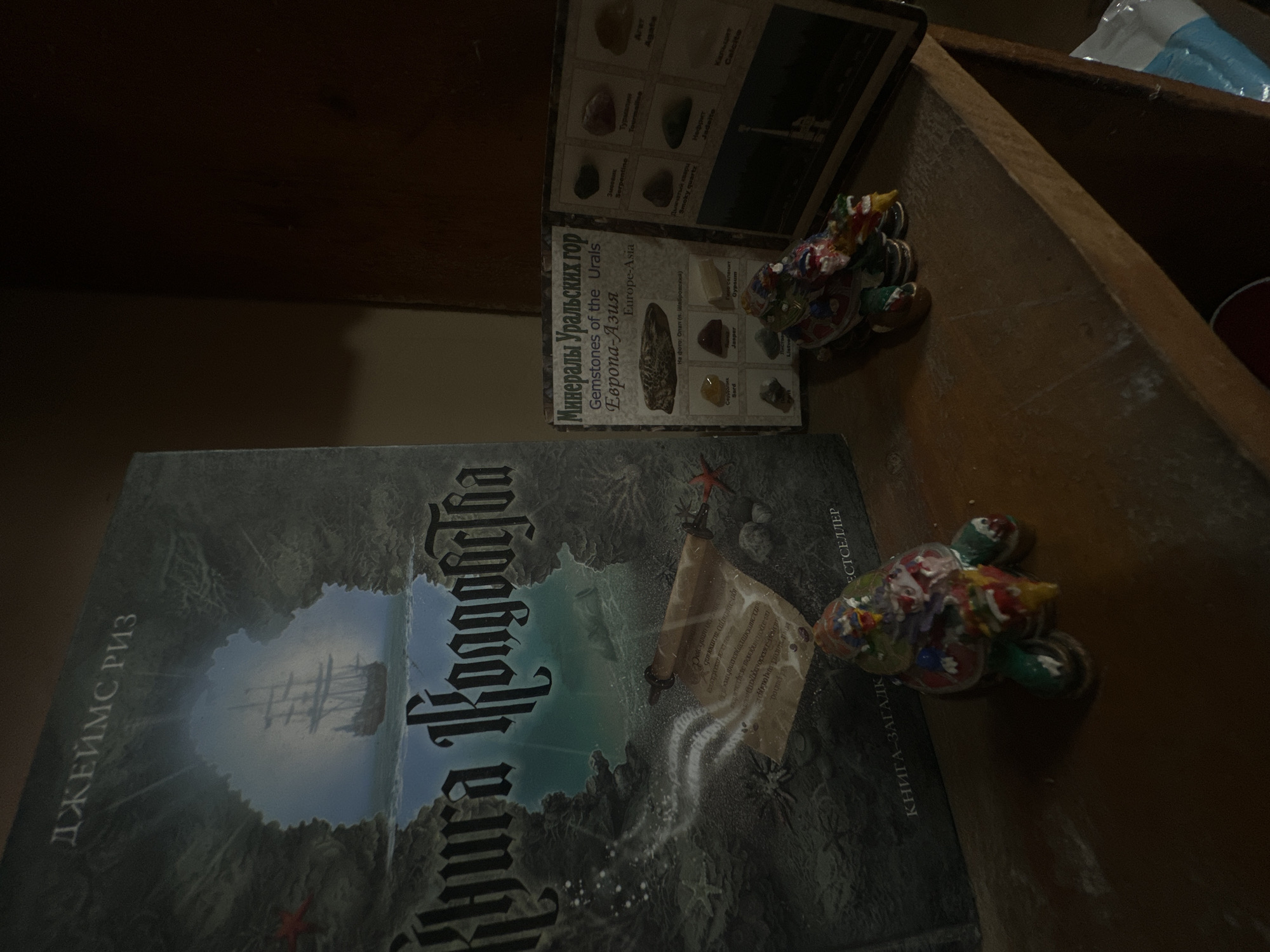 The image shows a dark wooden surface with several objects. In the foreground, a large book with a dark, textured cover is laid open. The cover features an illustration of a sailing ship silhouetted against a misty sky, surrounded by dark, rocky terrain. The book's title is visible in large, ornate font, partially obscured. To the right of the book, there is a smaller, open pamphlet or booklet displaying various mineral samples. The text on the booklet is in both Russian and English, identifying the minerals as "Gemstones of the Urals" from "Europe-Asia." Two small, colorful, dragon-like figurines are placed on the surface, one near the booklet and another further to the right. The lighting is dim, suggesting an indoor setting, possibly a shelf or table in a dimly lit room. The overall scene conveys a sense of collected items, perhaps a personal display of books and curiosities. There are no people visible in the image.