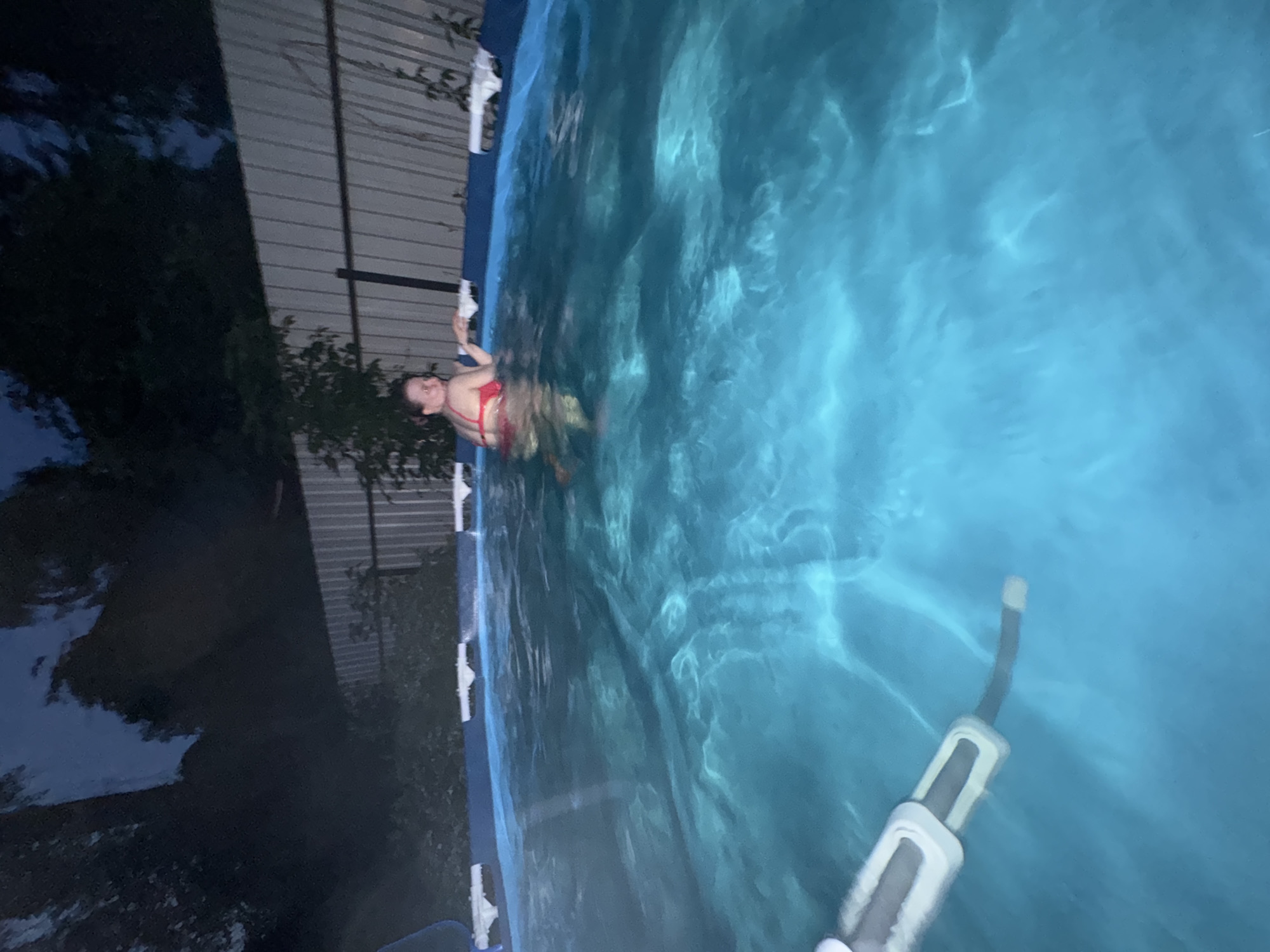 A woman in a red bikini is in a swimming pool at night. The pool is illuminated from within, casting a blue glow on the water. A fence and trees are visible in the background. The scene appears to be a residential backyard. There is no indication of the location being Moscow, Russia.