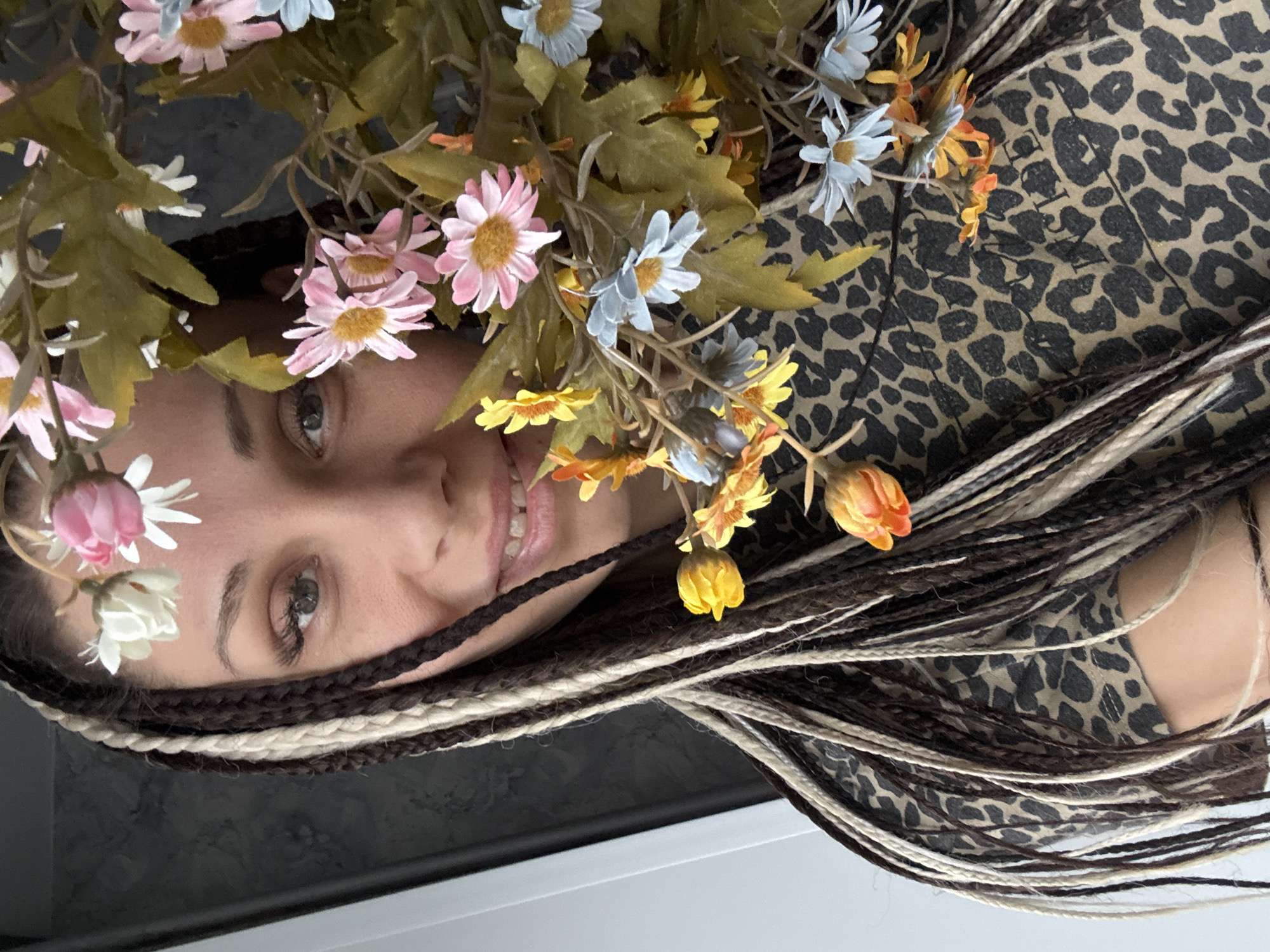 A woman is lying on a surface with a leopard print fabric covering part of her body. Her head is adorned with an arrangement of daisies in shades of pink, blue, yellow, orange, and white, intermixed with green leaves. She has braided hair in dark brown and blonde shades. The woman's face is visible, showing fair skin, light-colored eyes, and a slight smile. The background is a combination of black and white surfaces, suggesting an indoor setting. The location is not discernable from the image.