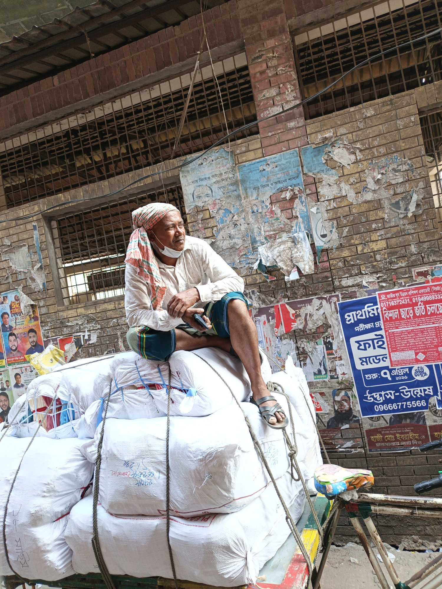 A man wearing a white shirt, a "lungi" (a traditional garment worn around the waist), and a colorful turban is sitting on top of a large pile of white bags tied together with ropes, on a rickshaw parked by a brick wall covered in posters. He has a medical mask hanging loose on his face, and he is holding a mobile phone in his hand. The background shows a building with barred windows. The Bangla script on the posters indicates the location is likely in Dhaka, Bangladesh. The scene suggests he may be a delivery man resting on his load.