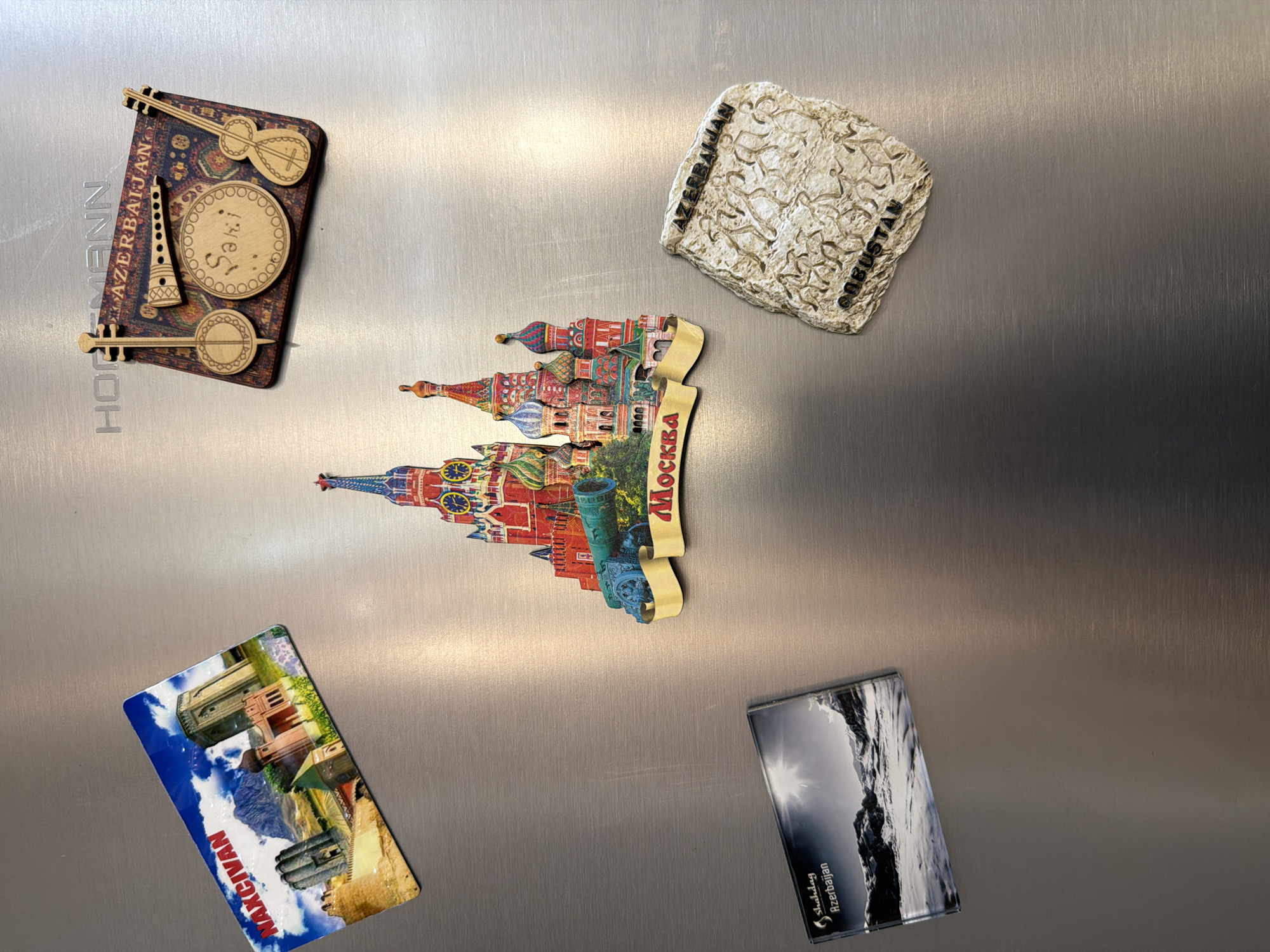 The image displays several magnets on a stainless steel refrigerator door. In the top left corner is a rectangular magnet featuring a colorful image of buildings and mountains with the text "NAXCIVAN" prominently displayed. To the right of this magnet is a wooden magnet shaped like a rug, adorned with carved musical instruments and the text "AZERBAIJAN" and "Saki". Below the wooden magnet is a vibrant, three-dimensional magnet depicting Saint Basil's Cathedral in Moscow, Russia, with the word "MOSKVA" on a banner. In the bottom left of the image, a horizontal magnet showcases a black and white landscape of snowy mountains and the text "Shahdag Azerbaijan". Finally, in the bottom right, a rough, stone-like magnet displays carvings of human figures and the text "AZERBAIJAN" and "GOBUSTAN". The refrigerator door shows some light scratches and reflections from the surrounding environment. There are no people visible in the image, and no specific location in the city of Sabirabad, Azerbaijan is depicted.