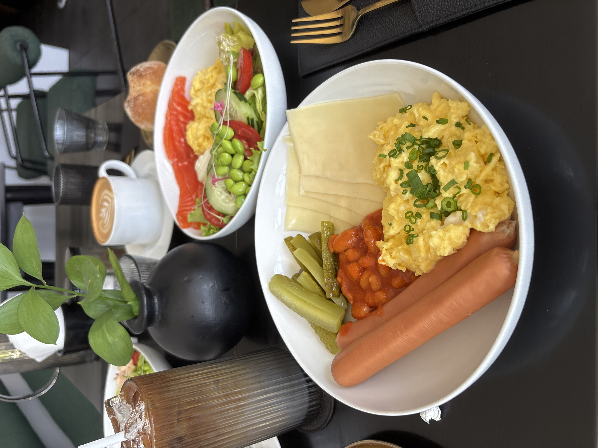 The image displays a vibrant breakfast or brunch spread laid out on a dark, reflective table, likely in a cafe or restaurant in Kuta Selatan, Indonesia. No people are visible.
Two main white bowls dominate the scene, each filled with various items. The bowl on the left contains a fresh salad mix with prominent slices of smoked salmon, fluffy scrambled eggs, vibrant green edamame pods, sliced cucumbers, and cherry tomatoes, garnished with small pink flowers. Adjacent to this bowl is a white cup of latte or cappuccino, adorned with intricate latte art.
The bowl on the right features a more traditional breakfast assortment, including scrambled eggs topped with chopped green onions, several slices of yellow cheese, a serving of baked beans in tomato sauce, sliced pickles, and two cooked hot dogs.
Other items on the table include a set of golden cutlery (fork and knife) resting on a black placemat to the upper right, and a small, black spherical vase holding green leafy stems, positioned to the left. A tall, ribbed glass containing an iced beverage with a white straw is visible in the lower-left foreground. A small pastry or bun is partially seen between the two main food bowls. The reflective tabletop creates blurred mirror images of the surroundings. The overall impression is a well-presented, appetizing meal, likely enjoyed during the morning or early afternoon. No visible text is present in the image.