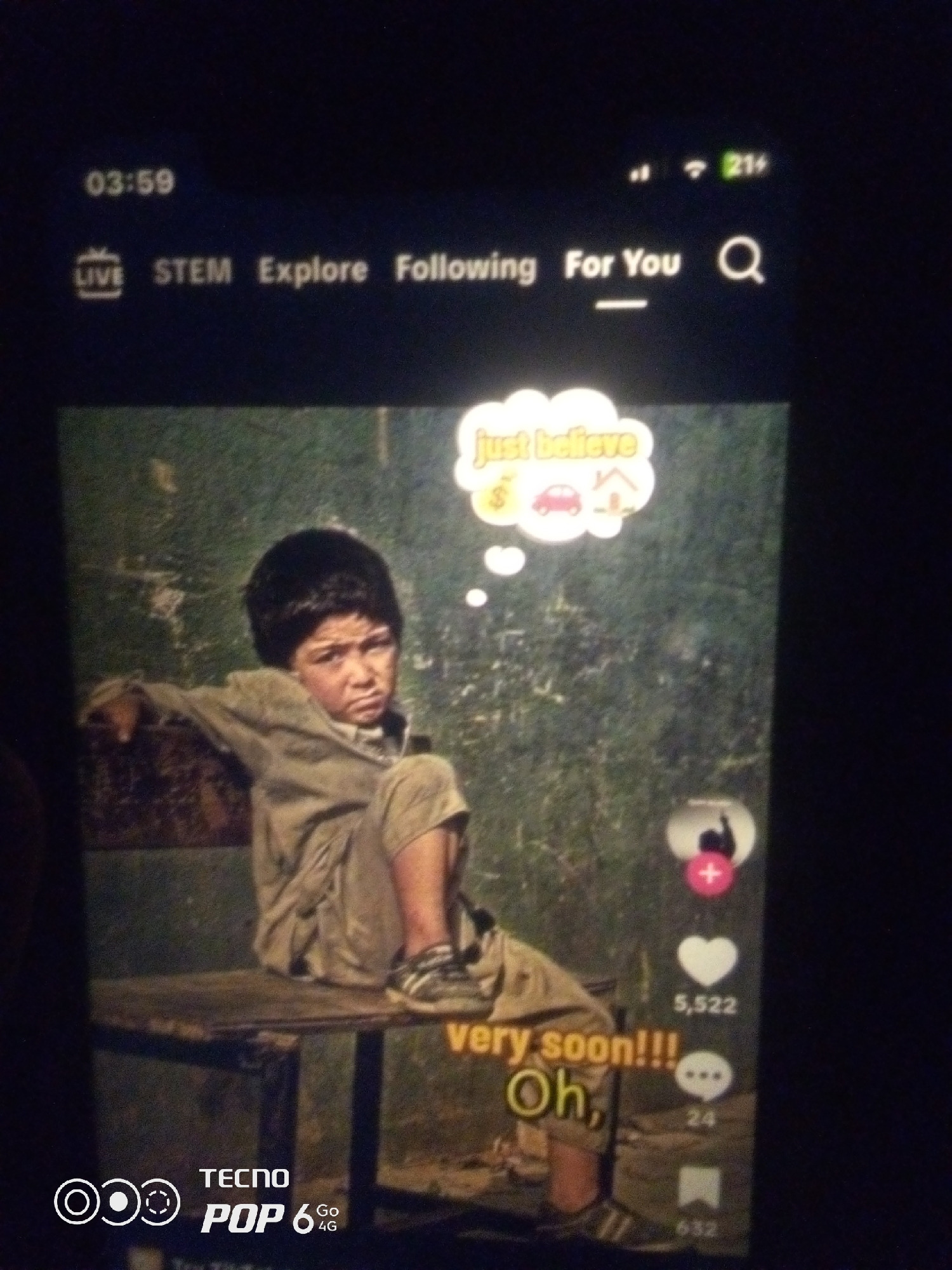 The image captures a young boy sitting on a wooden bench, looking directly at the camera with a pensive expression. He is wearing a light brown, oversized shirt and trousers, and appears to be barefoot. A thought bubble floats above his head, containing a dollar sign, a car, and a house icon, suggesting a wish for financial stability, transportation, and shelter. The background is a blurred, textured green surface, possibly a wall or foliage, with some lighter patches. On the right side of the image, a social media interface is visible, with icons for likes, comments, and shares, and corresponding numbers: 5,522 likes and 24 comments. The bottom left corner displays "TECNO POP 6 4G" branding. The image quality is low, suggesting it was taken with a mobile phone in low light conditions. The image is not geotagged, and there is no information to confirm or deny its location in Howayi, Nigeria.