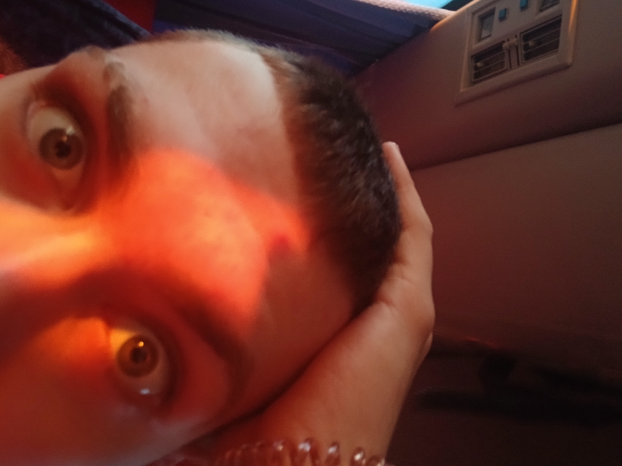A close-up selfie shows a person's face illuminated by an orange light, casting a warm glow on their features. The person has a short haircut and light skin. Their eyes are wide, with visible brown irises. The person's hand is visible on the right side of the frame, resting against their head. Behind them, a panel with vent-like openings is visible. The image does not contain enough information to confirm it was taken in Ustinovo, Russia.