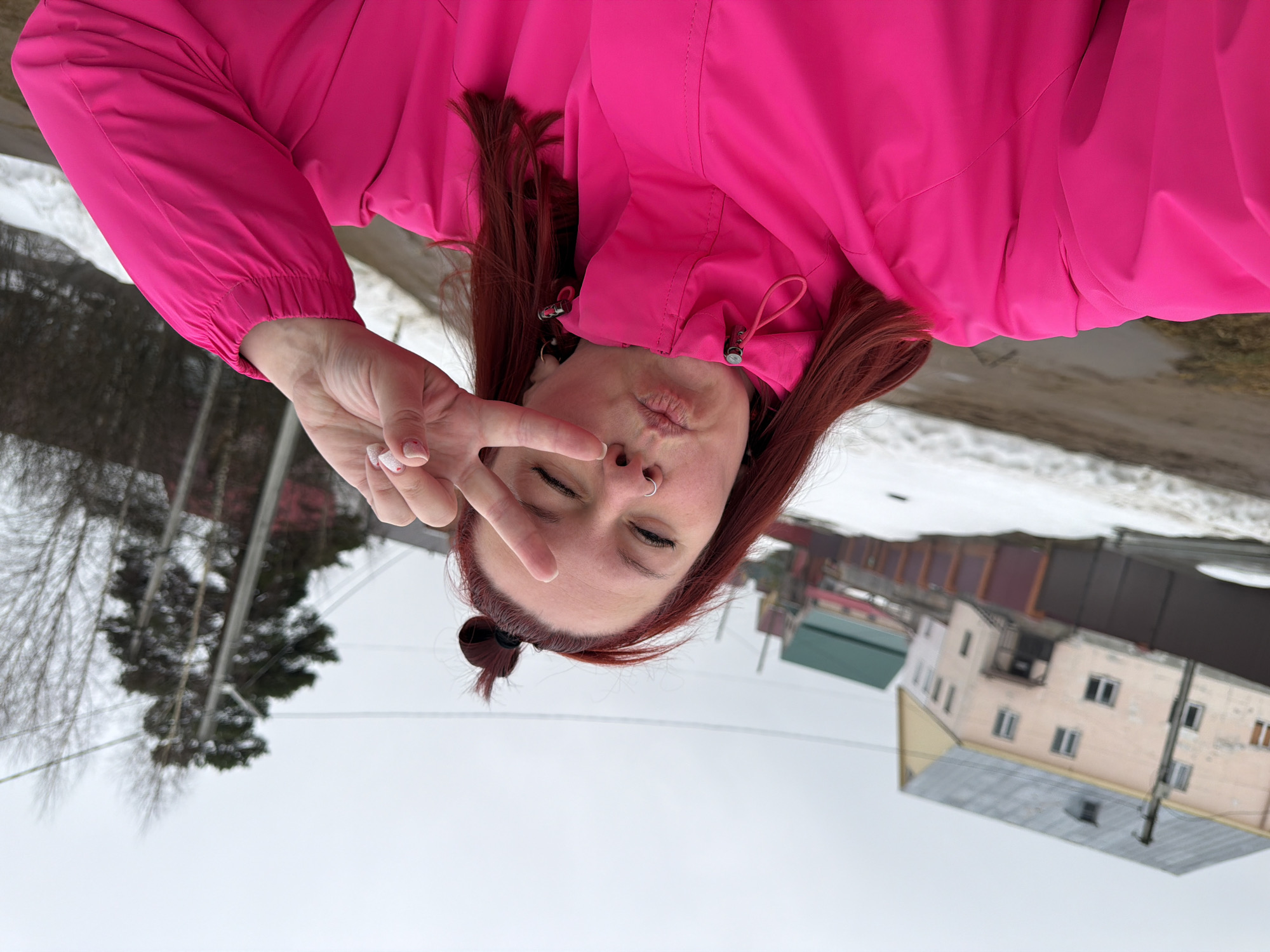 A woman wearing a bright pink jacket poses for a selfie in Chaikovsky, Russia. She is making a "peace" sign with her left hand while puckering her lips. The woman has red hair pulled into a small bun on top of her head and a nose ring. Behind her are snow-covered ground, trees, buildings, and overhead wires, characteristic of an urban environment in Russia. The photo is taken from a low angle and rotated 180 degrees.