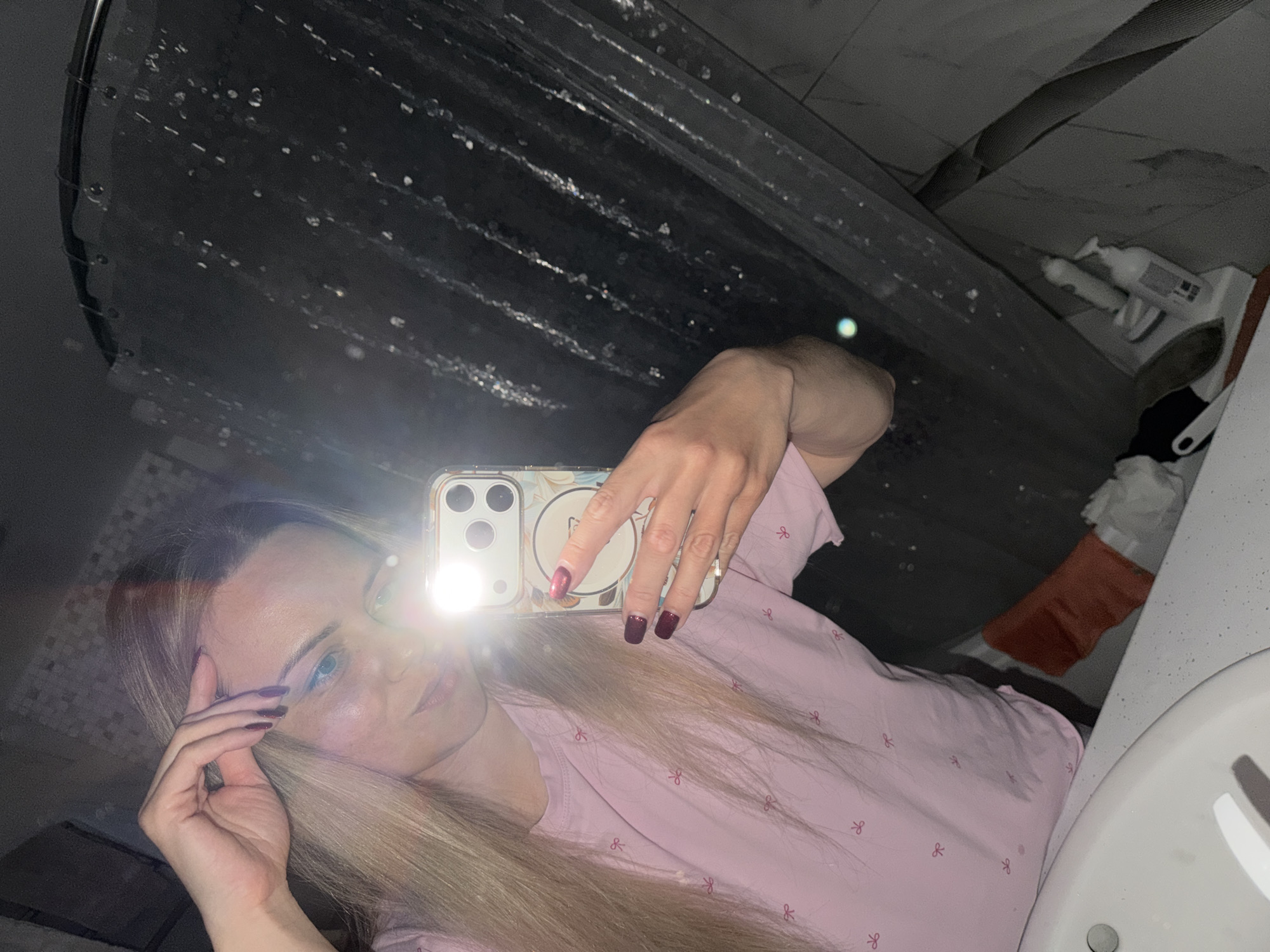 A person with light blonde hair and blue eyes takes a selfie in a bathroom. The person wears a light pink top with a bow pattern. The right hand holds a smartphone, obscuring part of the face with the phone's light. The left hand is raised to the forehead. The background includes a shower curtain, bathroom sink, and some toiletries. The location is a bathroom, but there is no information to place it in Votkinsk, Russia.