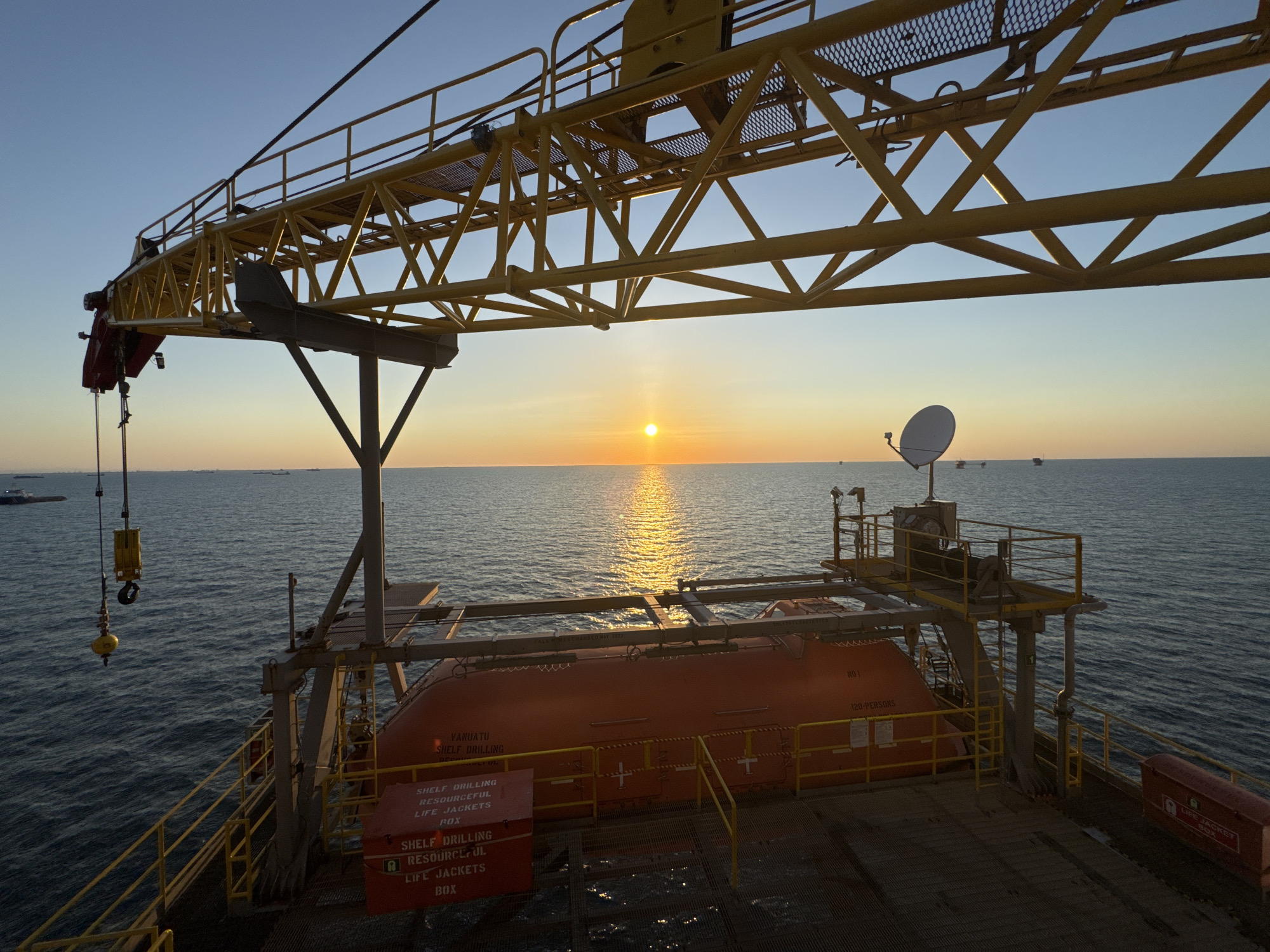 The image shows an offshore drilling rig at sunrise. The sun is low on the horizon, casting a warm glow across the water. A large yellow crane extends from the top left of the frame, with its boom angled upwards. Below the crane is a bright orange lifeboat, with "VANUATU SHELF DRILLING" printed on its side. The lifeboat is mounted on a platform with yellow railings. A red life jacket box is visible on the right side of the platform. The ocean is calm, with small waves. In the distance, several other ships and oil rigs can be seen. The sky is clear and blue, transitioning to a soft orange and yellow hue near the horizon. The location is an offshore oil rig, not in a city.