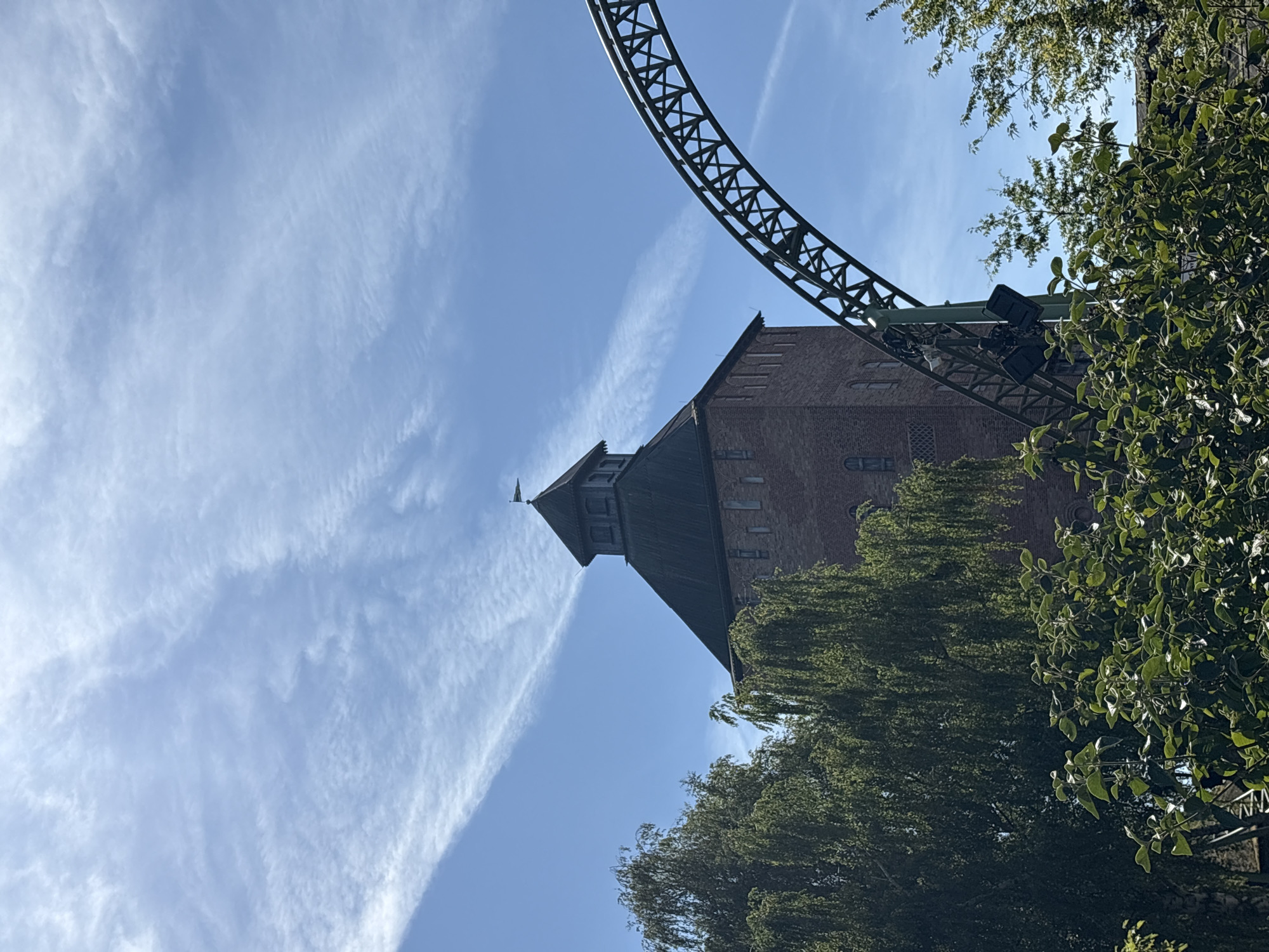 The image shows a brick tower with a dark roof and a small flag on top, set against a bright blue sky with wispy white clouds. In the foreground, lush green trees with long, drooping branches partially obscure the tower. To the right of the tower, a section of a green roller coaster track curves upwards, also partially visible through the foliage. The scene appears to be a sunny day at an amusement park, likely the Hansa-Park in Sierksdorf, Germany, given the characteristic architecture and presence of a roller coaster. No people are visible in the image.