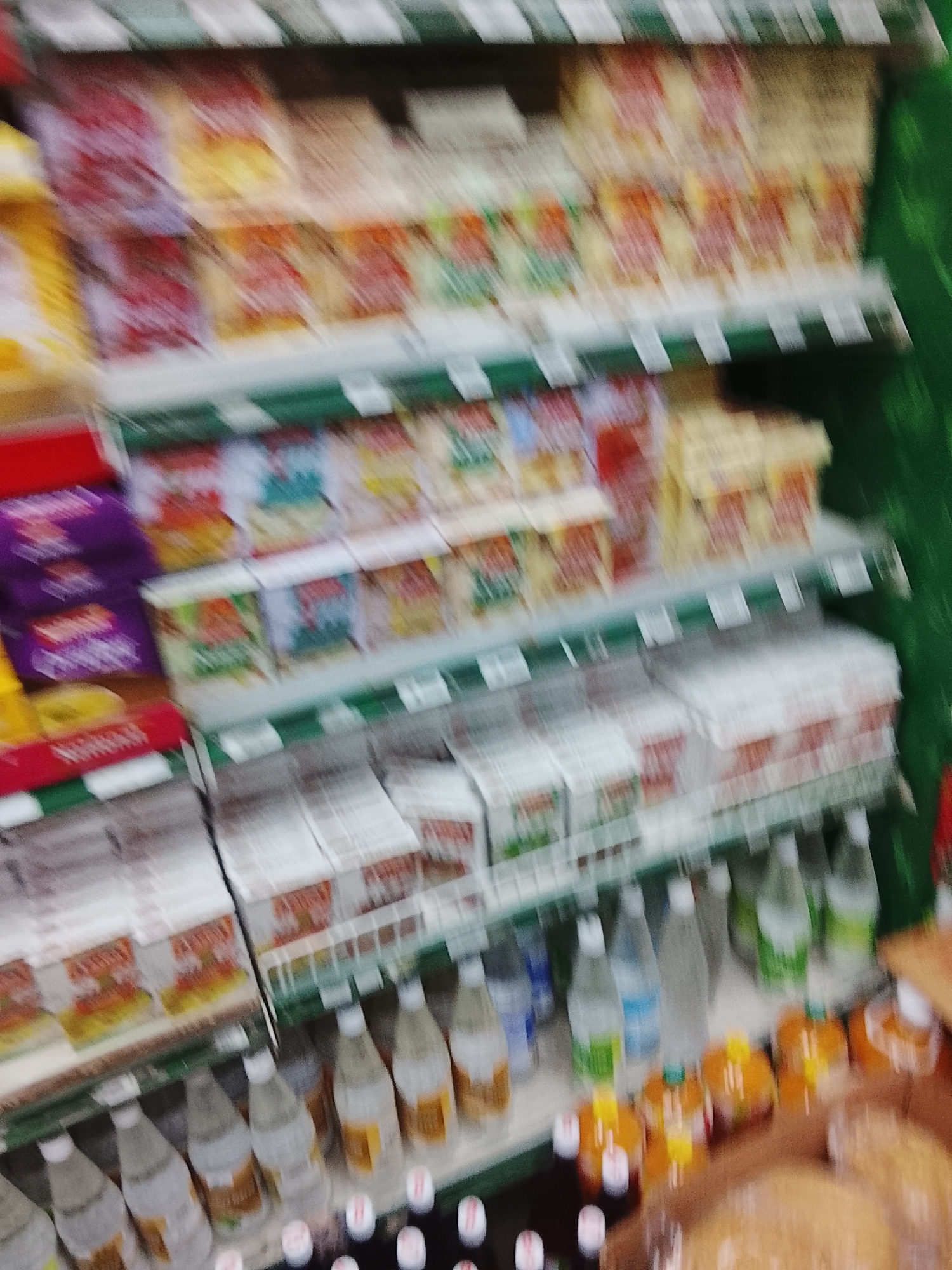 The image shows shelves stocked with various food and beverage items in what appears to be a grocery store. The shelves are arranged in tiers, and the products include items in boxes, bottles, and jars. The items on the shelves are not clearly discernible due to the image's blurriness. There is no clear indication of the exact location within Gulgasht Colony, Pakistan. No people, subjects, or actions can be identified with certainty. The environment consists of the interior of the store with shelved products.