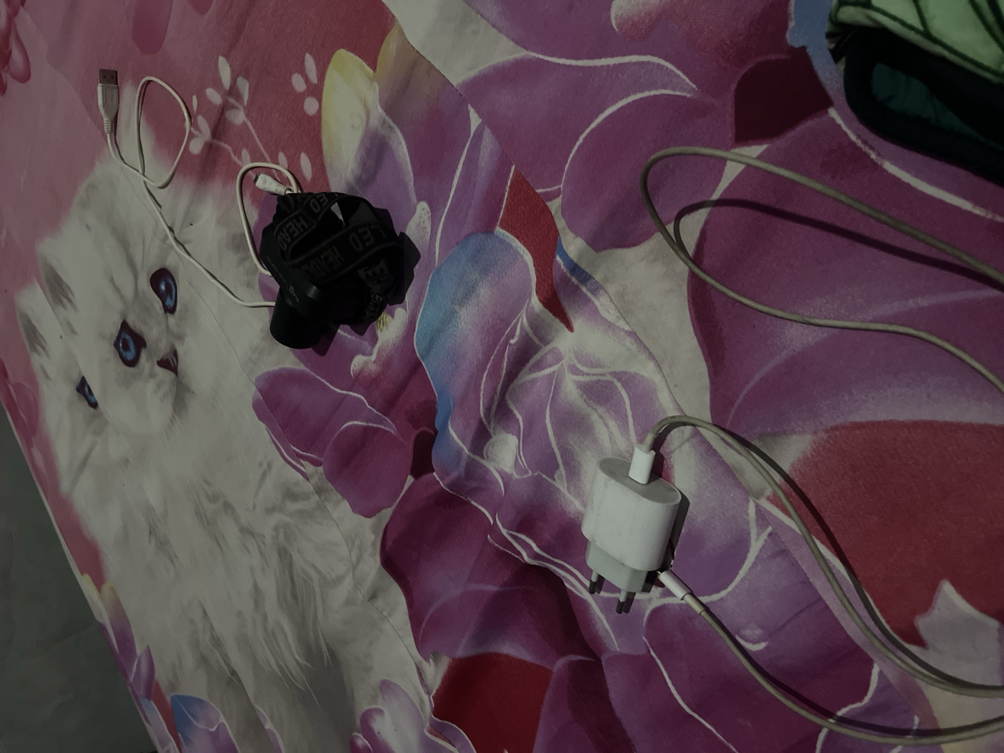 The image shows a close-up of a bed with a pink floral patterned duvet that has a white cat design on it. On the duvet, there are two electronic charging devices. One is a white USB wall adapter plugged into a white cable. The other is a black LED headlamp with its strap visible, also connected to a white cable with a USB connection. The scene is indoors, likely a bedroom, given the bed and the soft lighting. There are no people present in the image. The location is not identifiable as Kebumen, Indonesia, or any specific city.